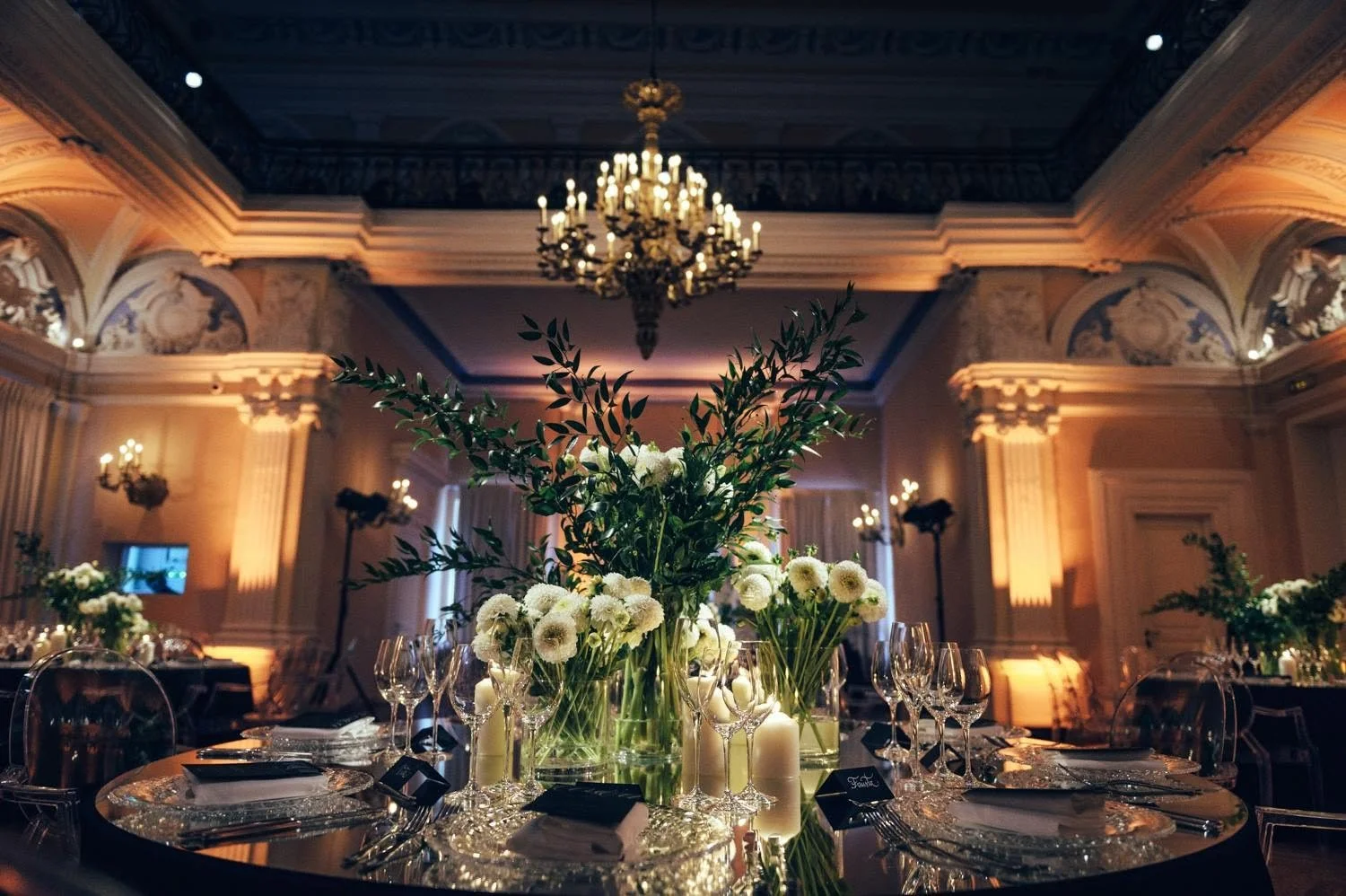 Elegant banquet hall with a large floral centerpiece of white flowers and greenery on a polished table, surrounded by glassware, candles, and place settings in a luxurious room with chandeliers and ornate architectural details.