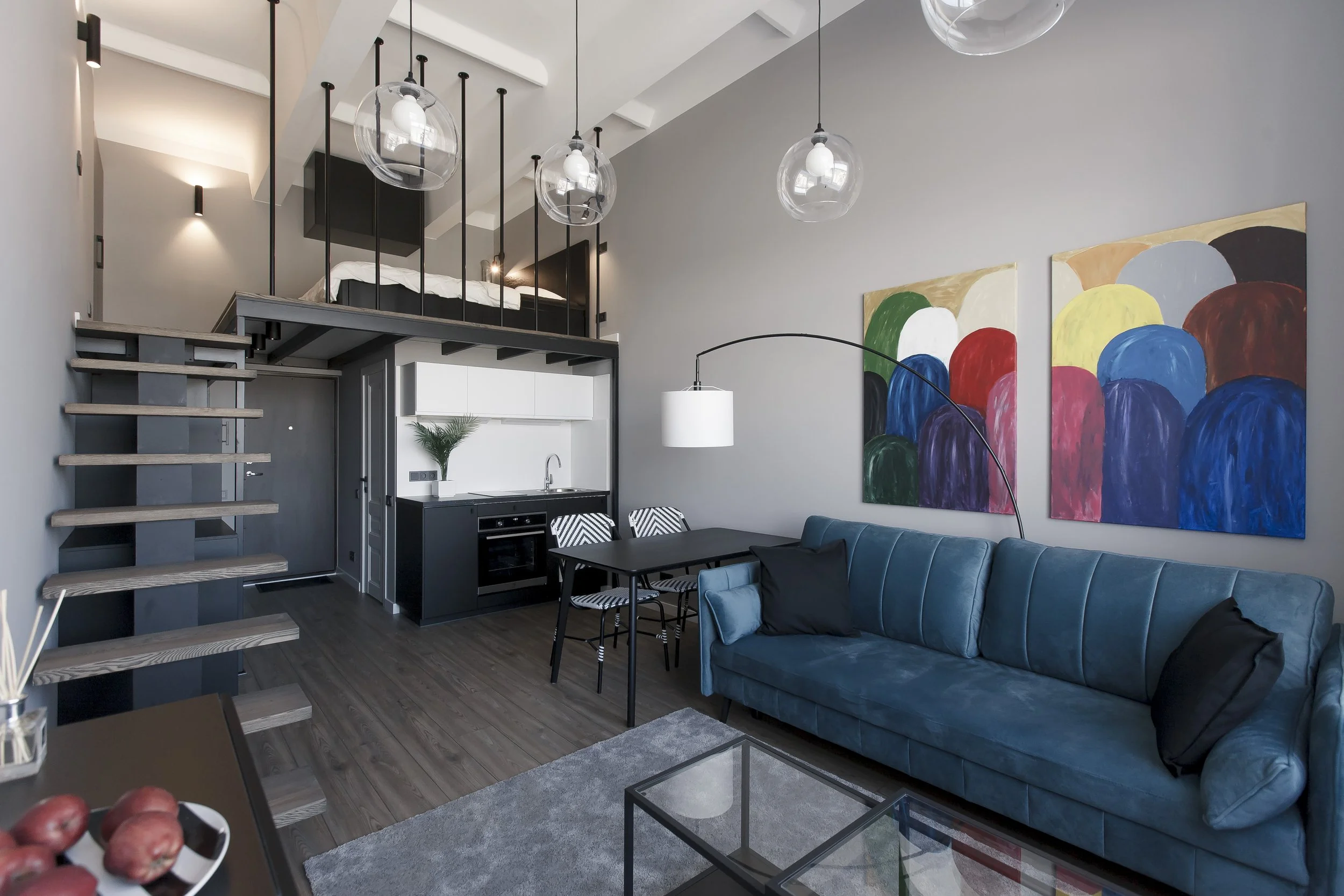 Modern living room with a high ceiling, featuring colorful abstract wall art, a blue velvet sofa, a black dining table with chairs, a loft bed accessed by stairs, pendant and wall lighting, and a small kitchen area.