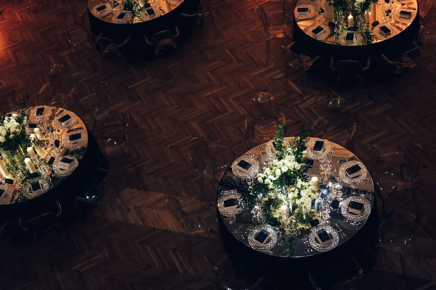 Elegant banquet tables with floral centerpieces, candles, glasses, plates, and silverware set up for an event on a dark wooden floor.