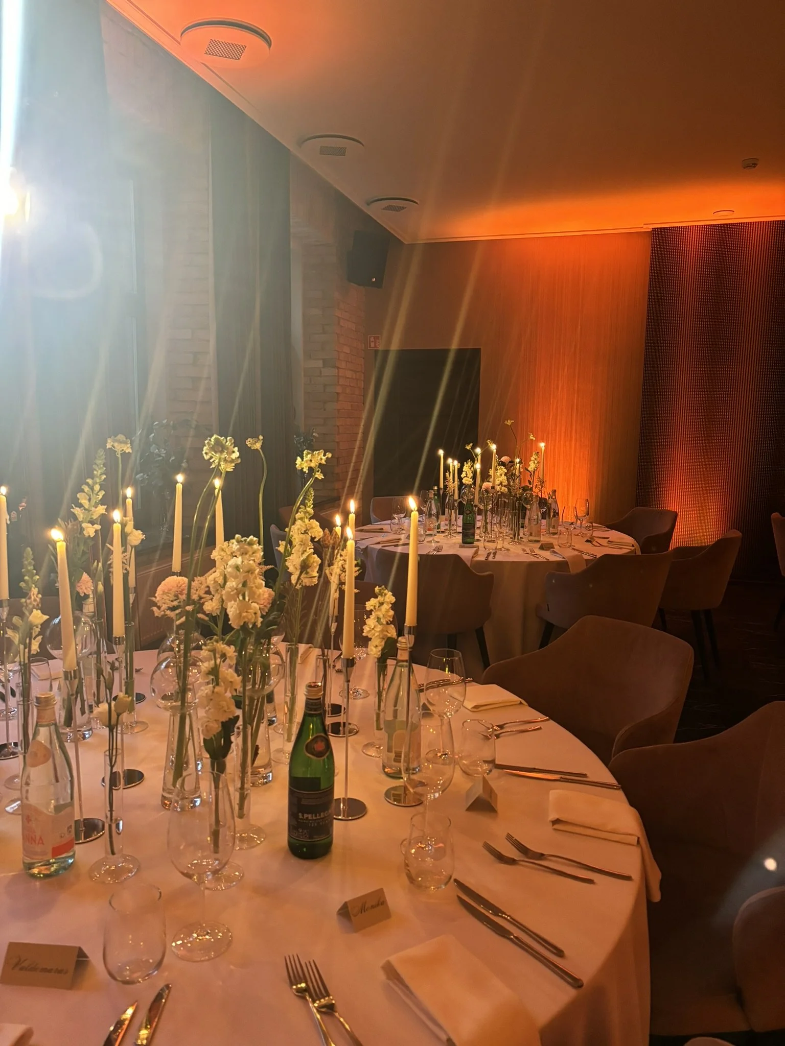 A restaurant table set for a formal dinner with white tablecloth, floral centerpieces, candles, wine glasses, cutlery, and place cards, in a warmly lit interior with wooden paneling and chairs around the table.