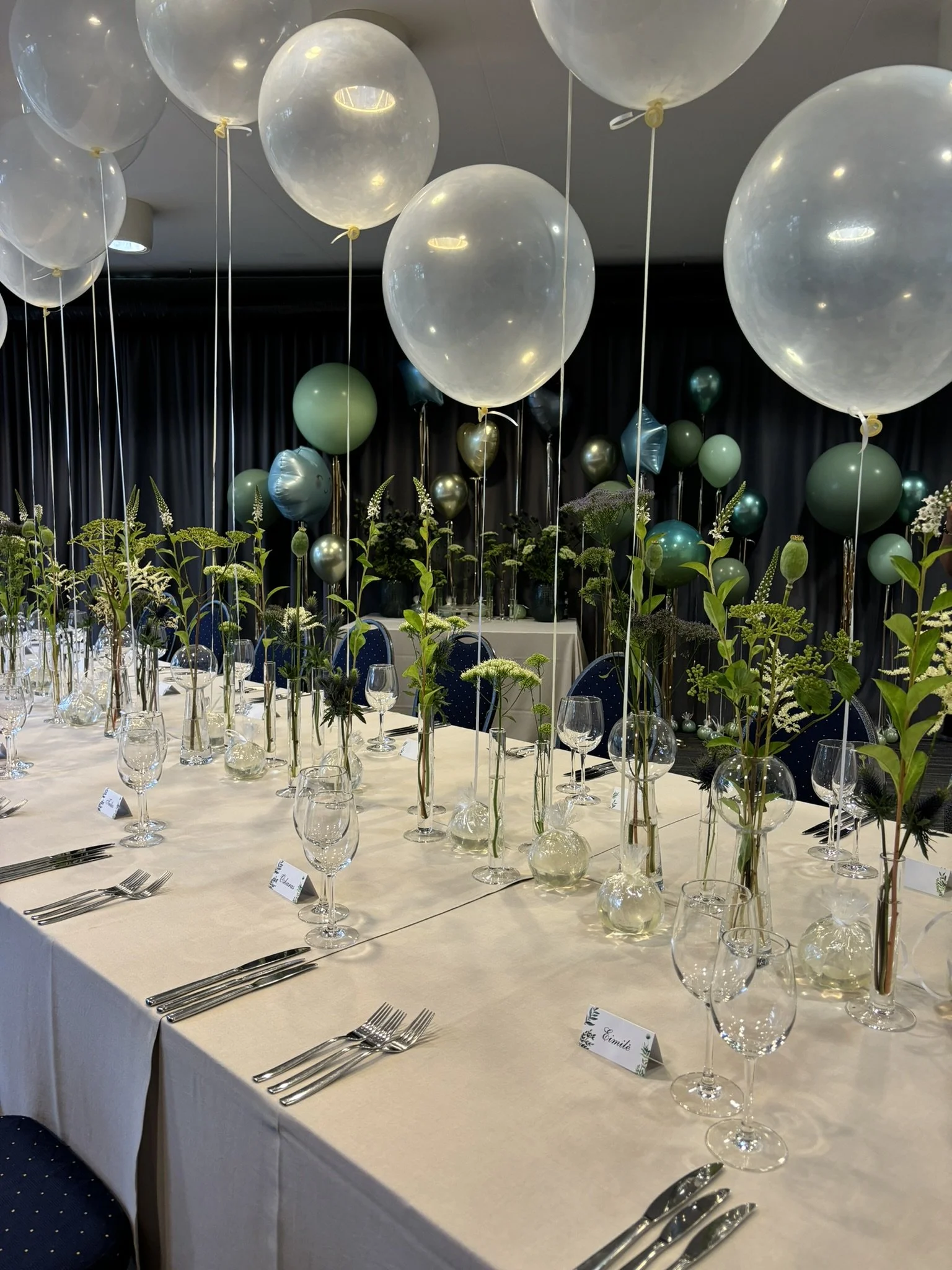 Table decorated with numerous helium balloons in various shades of white, green, and metallic, with floral centerpieces and place cards, set for a formal event or celebration.