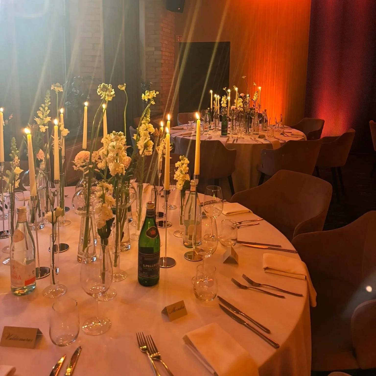 Elegantly set dining table with white tablecloth, tall yellow candles, floral centerpieces, wine glasses, bottles, and silverware, in a warmly lit room with orange lights and modern decor.