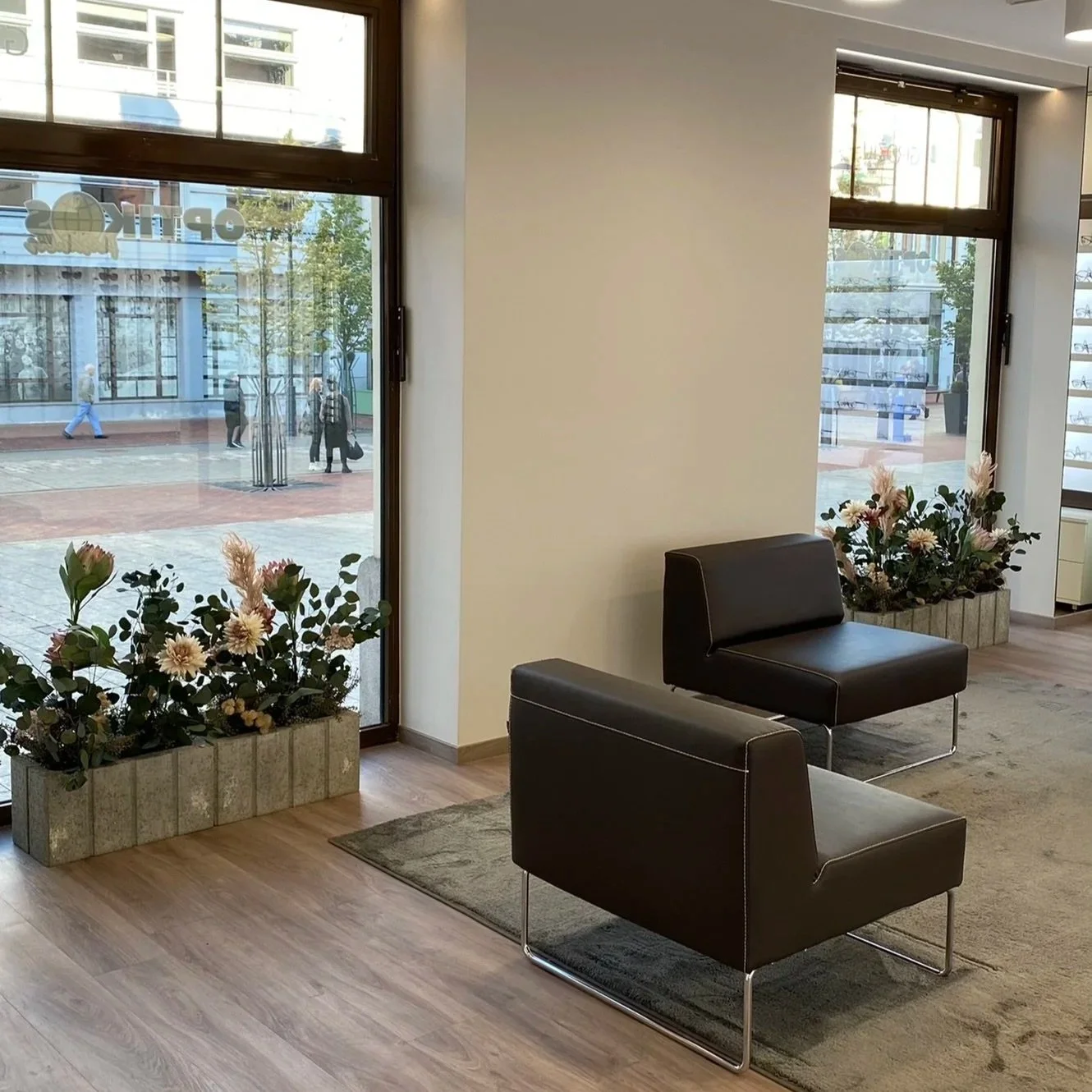 Interior view of a modern waiting area with black leather chairs, floral arrangements in rectangular planters, large windows overlooking a street with pedestrians, and a wooden floor.