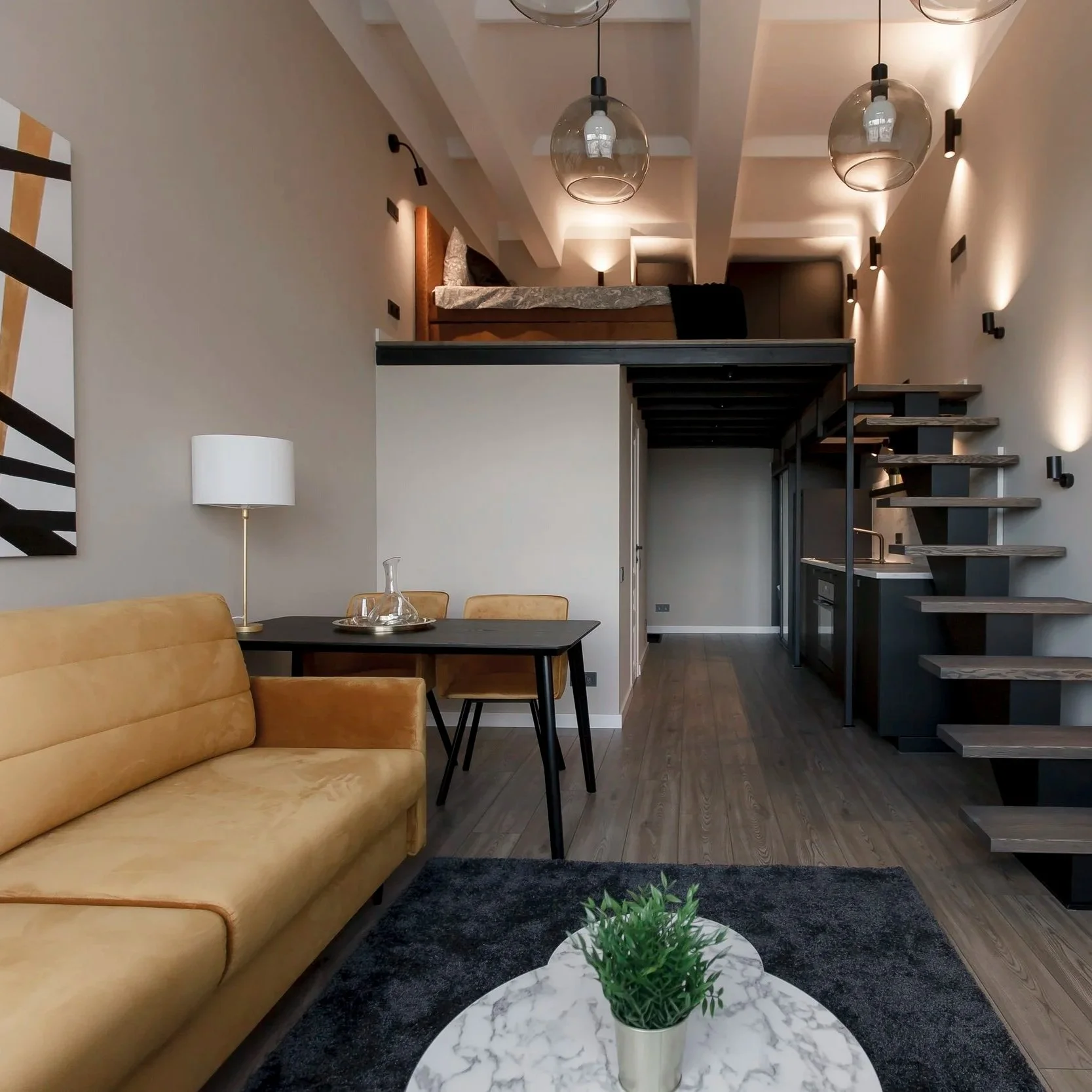 A modern open-concept living space featuring a tan sofa, a black table with a glass decanter, a white marble-topped coffee table with a green potted plant, a black kitchen island with a sink, and stairs leading to a loft area with a bed and hanging globe lights. The walls are painted white with various wall-mounted lights and artwork.