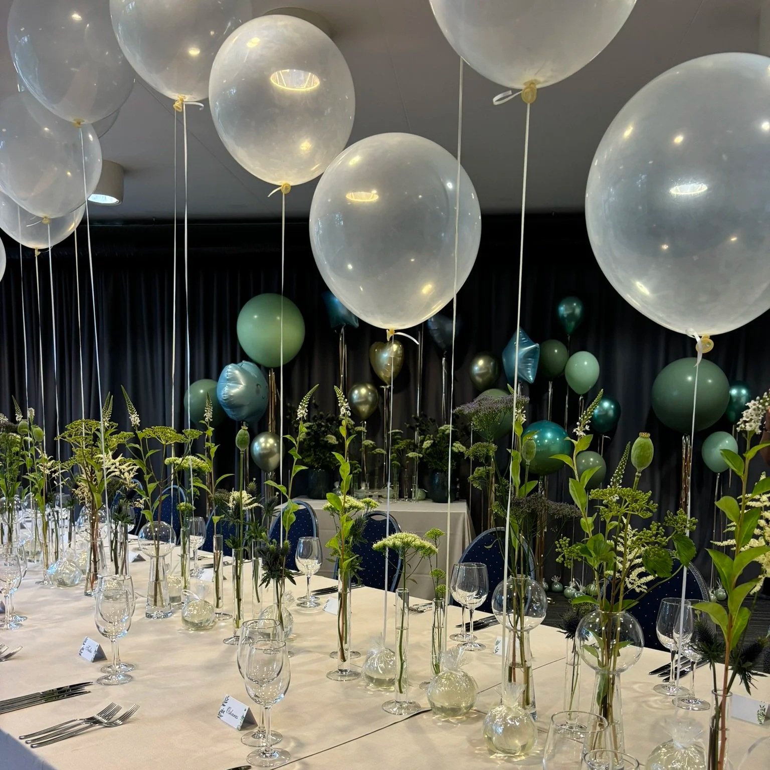 Decorative event table with clear and teal balloons floating above, decorated with various green plants and flowers in vases, with blue chairs and a dark curtain backdrop.