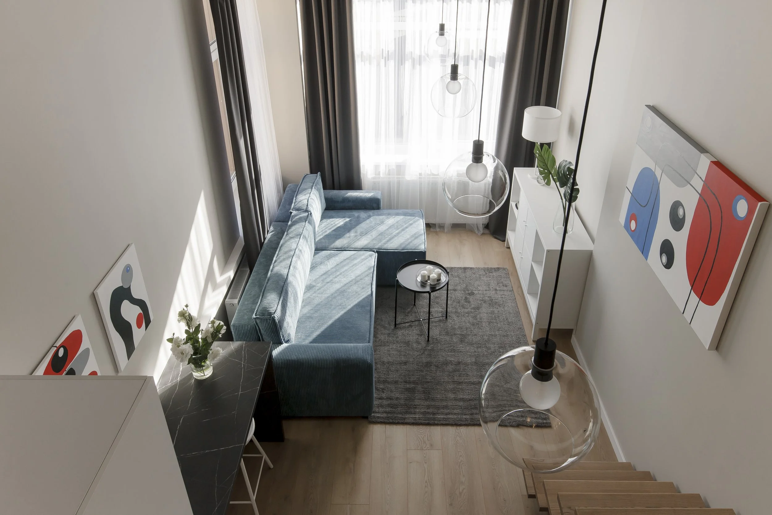 Living room with light-colored walls, a blue velvet sofa, a black marble side table with candles, a white cabinet, modern hanging globe pendant lights, artwork on the walls, a floor lamp, a flower vase, and large windows with dark curtains.