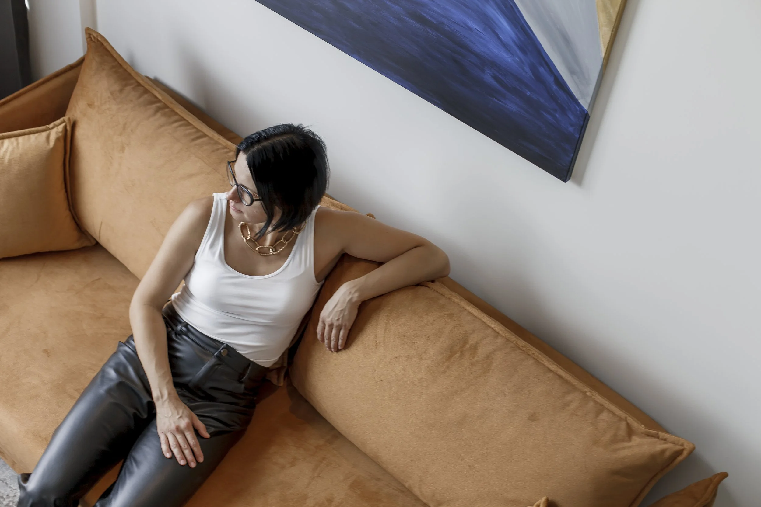 A woman with black hair and glasses sitting on an orange sofa, wearing a white top, black leather pants, and a gold chain necklace, with a blue abstract painting on the wall behind her.