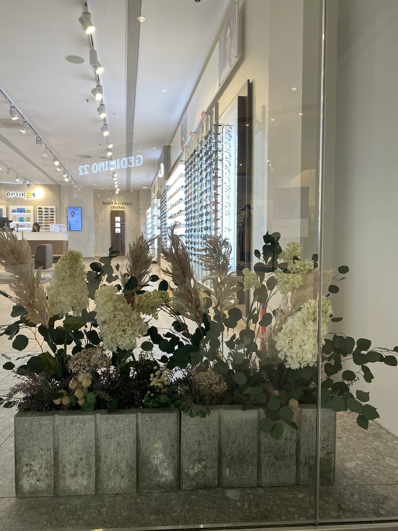A large flower arrangement with white hydrangeas, dried plants, and greenery in a concrete planter, positioned near a glass wall inside a modern store or mall.
