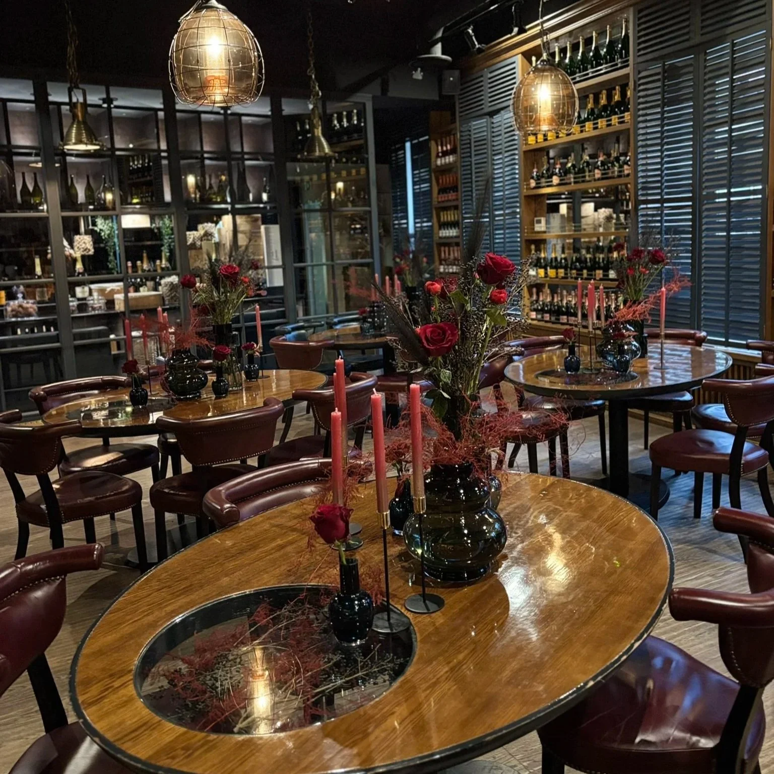 Interior of a restaurant with wooden tables decorated with black vases and red flowers, including roses, and pink taper candles. The background features shelves with wine bottles and a wall with blue shutters, illuminated by pendant lights.