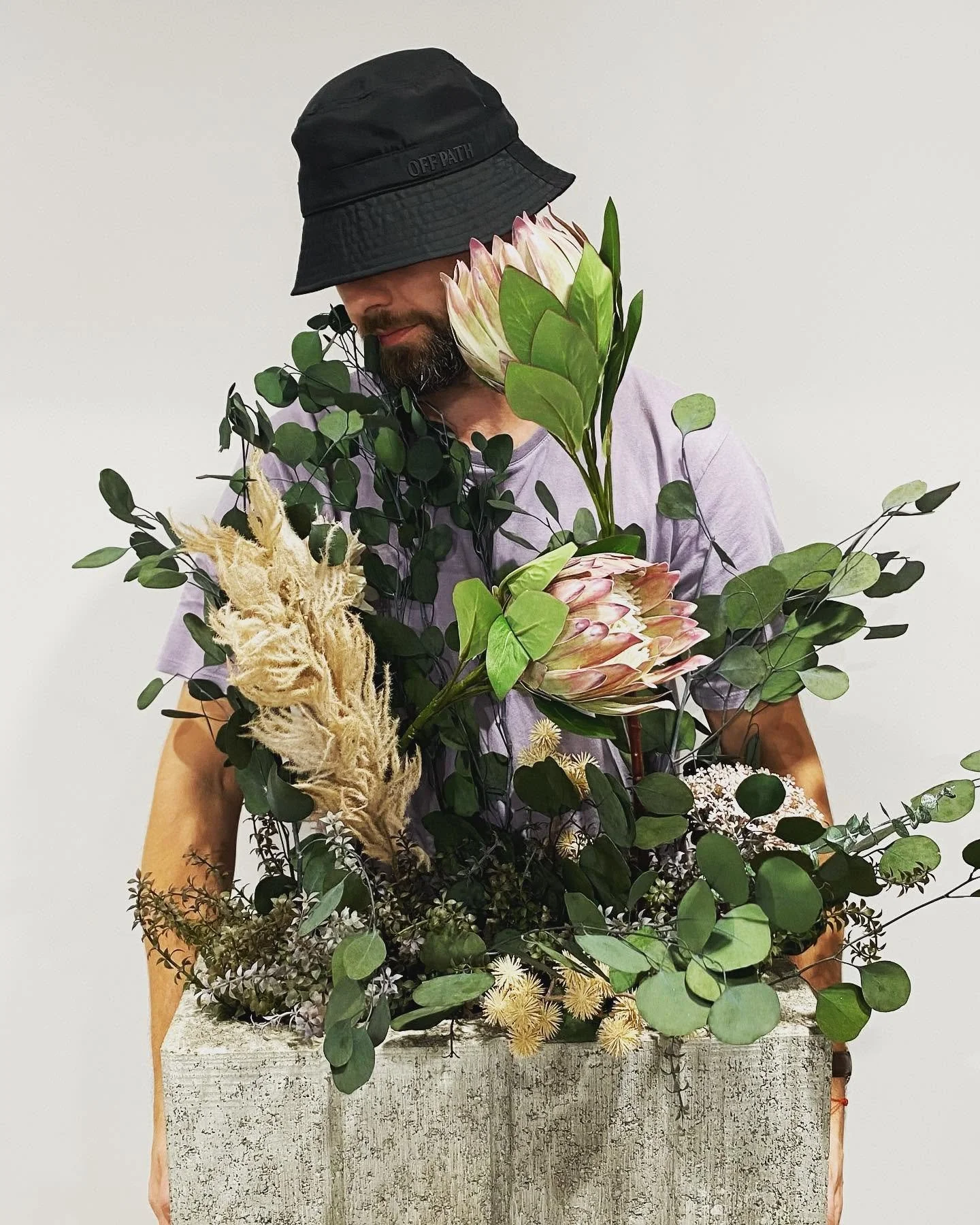 Man in a black bucket hat, purple shirt, holding a large arrangement of various flowers and green leaves against a plain white background.