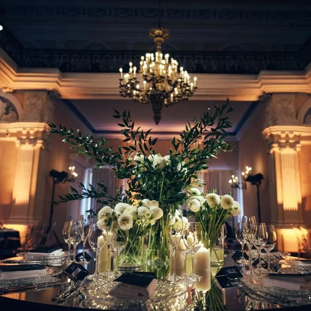 Elegant table setting with a large floral centerpiece, surrounded by glassware and candles, in a grand decorated hall with chandeliers.