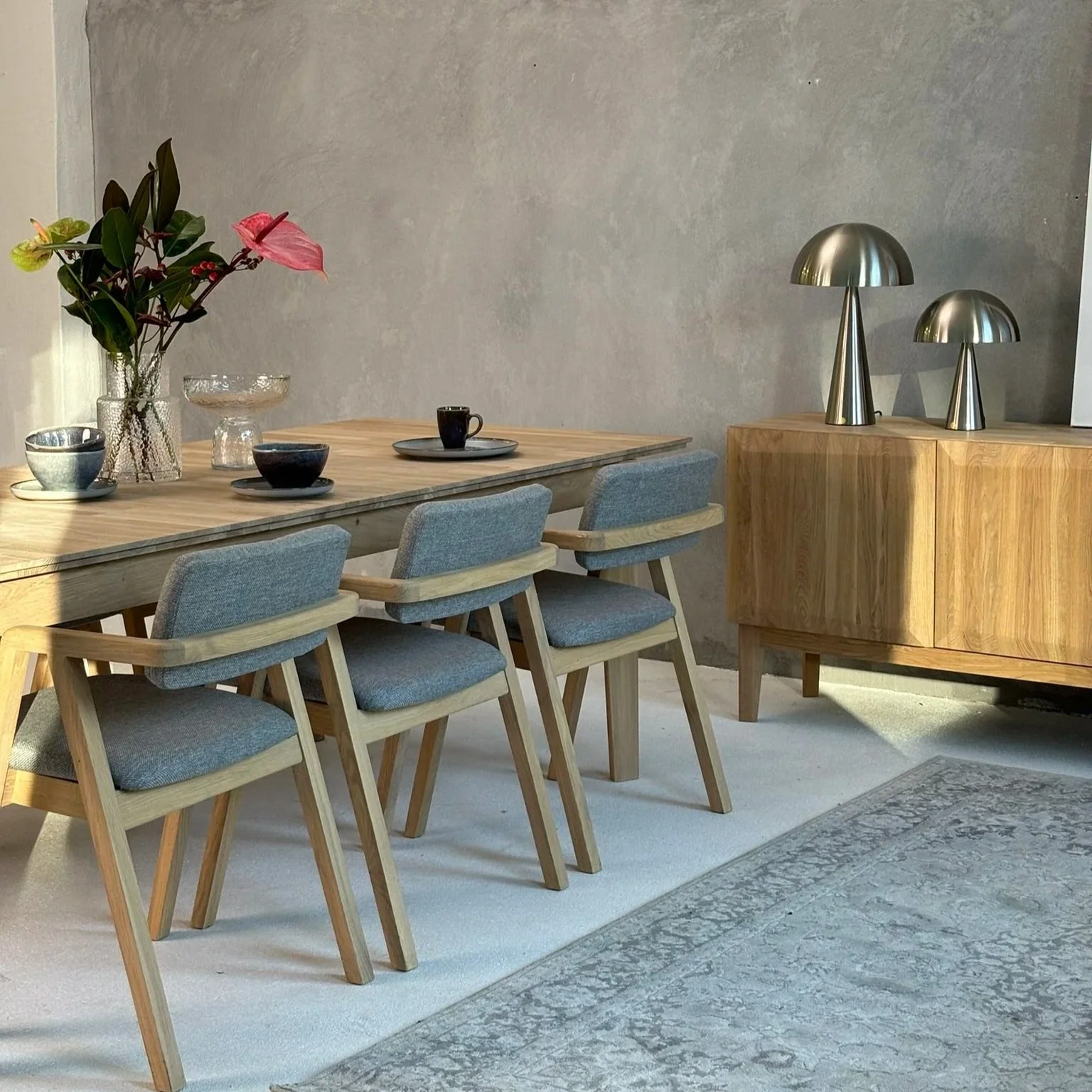 A dining room with a wooden table, four light blue upholstered chairs, a glass vase with pink and green flowers, several small bowls and cups on the table, a wooden sideboard with two metallic table lamps, a patterned rug, and a plain light-colored wall.