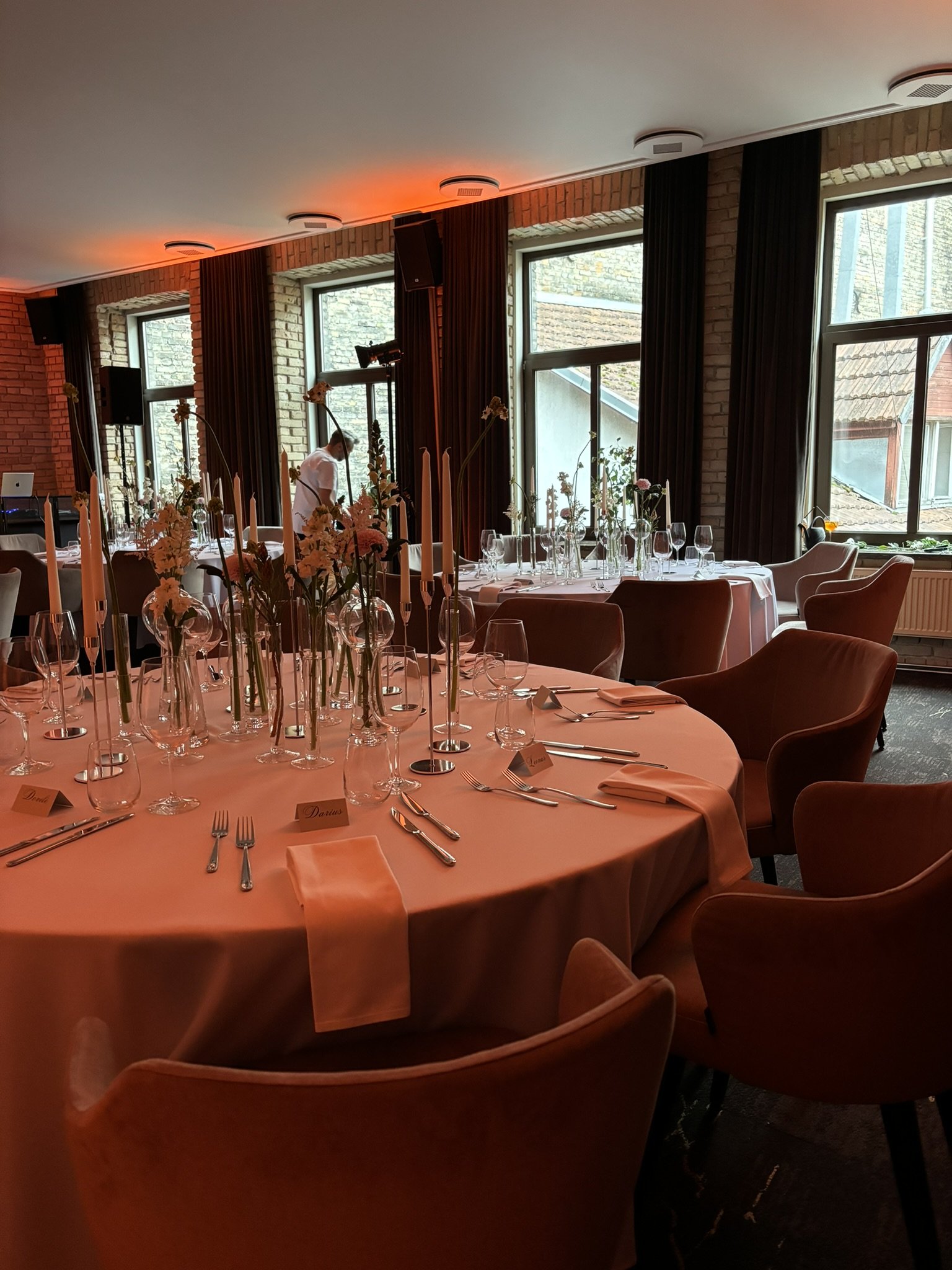 Indoor dining area with round tables covered in white tablecloths, set with wine glasses, silverware, and candles, with floral centerpieces. Large windows with black curtains let in natural light, and brick walls are visible.