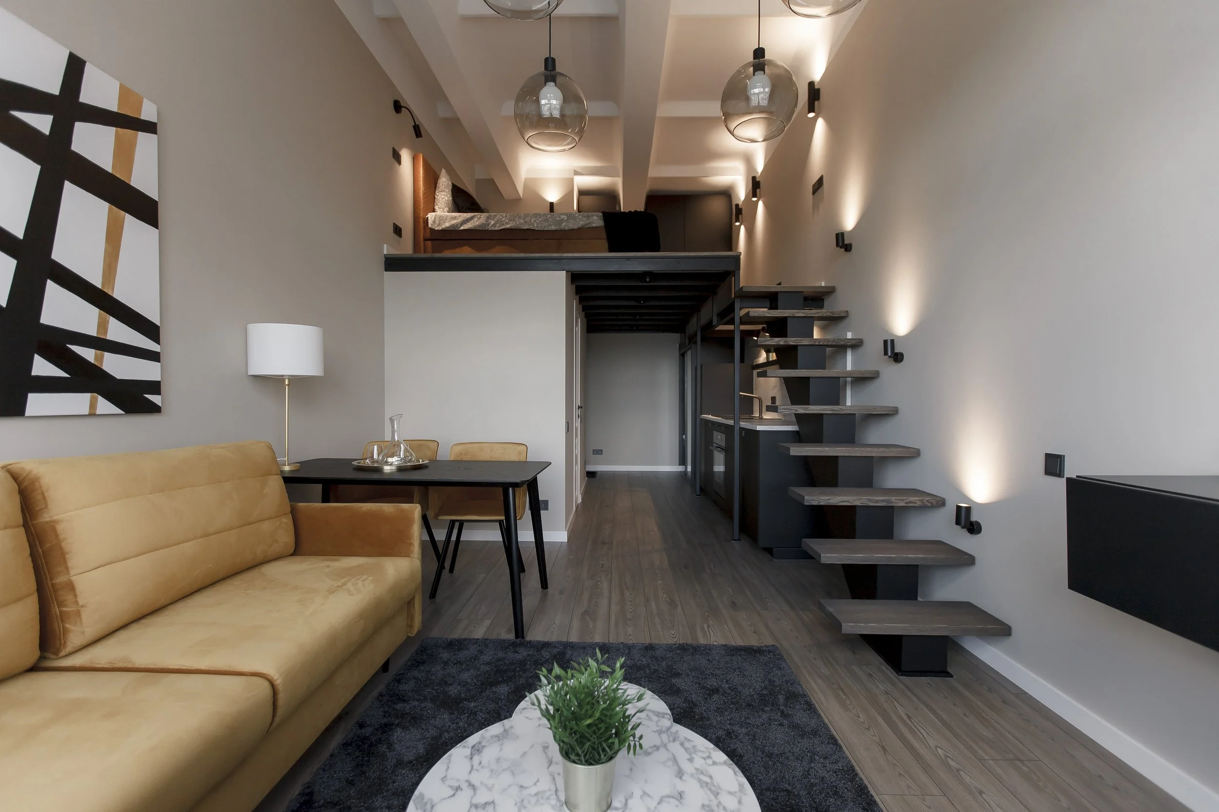 Living room with a beige couch, black coffee table with a potted plant, a yellow dining chair, a dining table, wall artwork, modern staircase, and loft with bed in the background. Modern lighting and wall sconces create a cozy atmosphere.