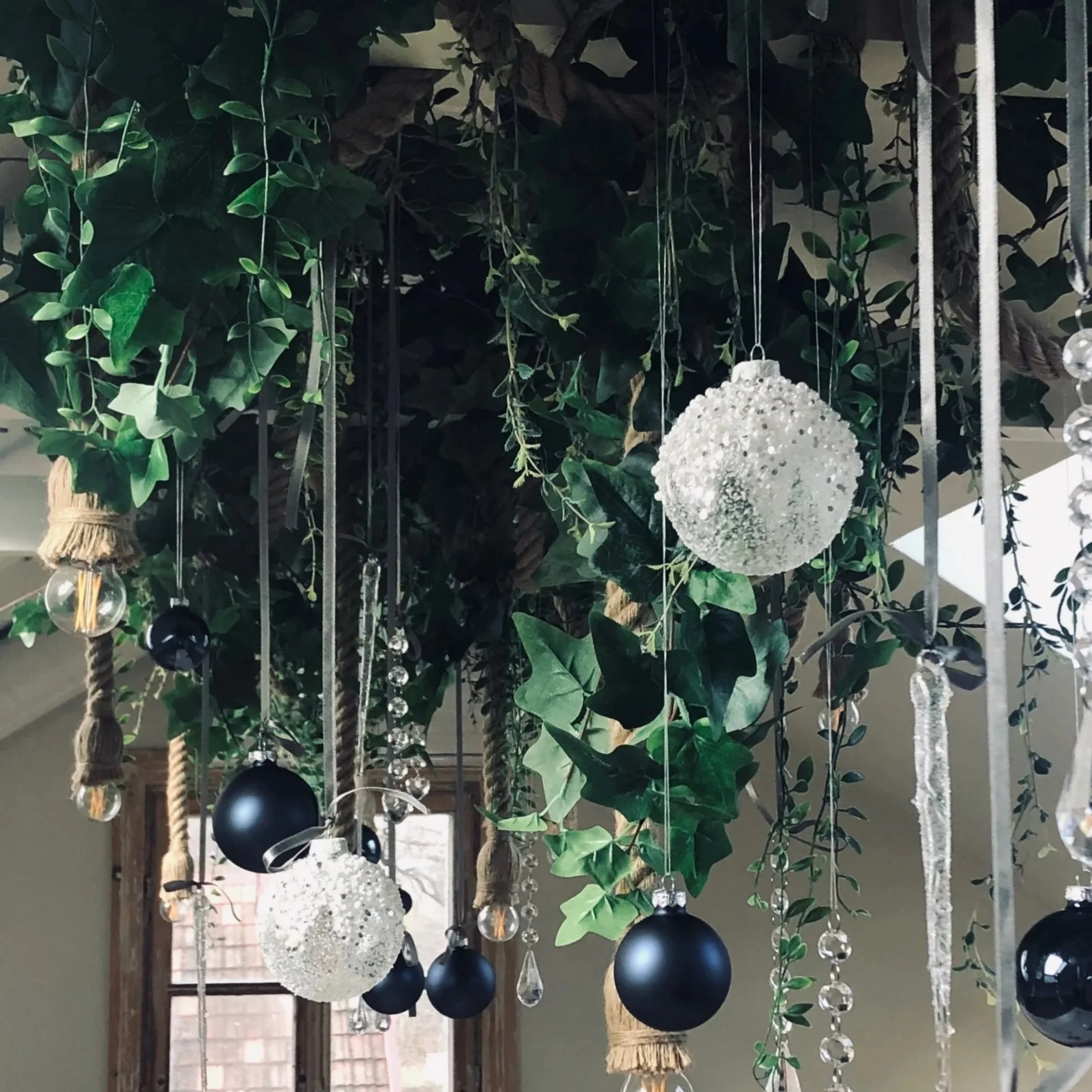 Decorative hanging ornament with black and white spherical objects, clear glass beads, and artificial green leaves, with a background of a porch and trees.
