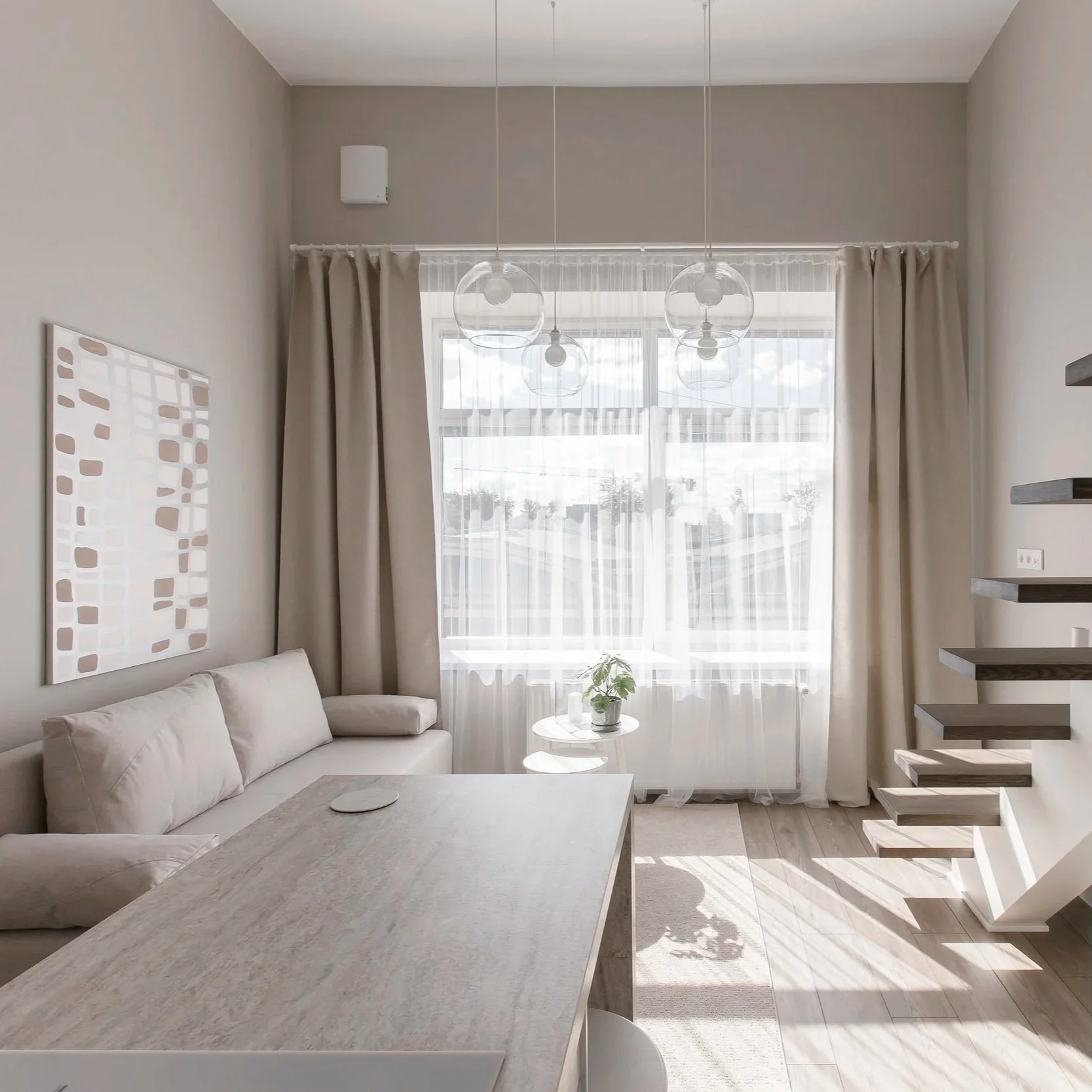 A bright, modern living room with beige walls, white curtains, and a large window allowing natural light. There is a white couch with cushions, two small round tables near the window with a small potted plant, a minimalist wall art piece, a set of floating shelves, and stylish staircase with floating steps.