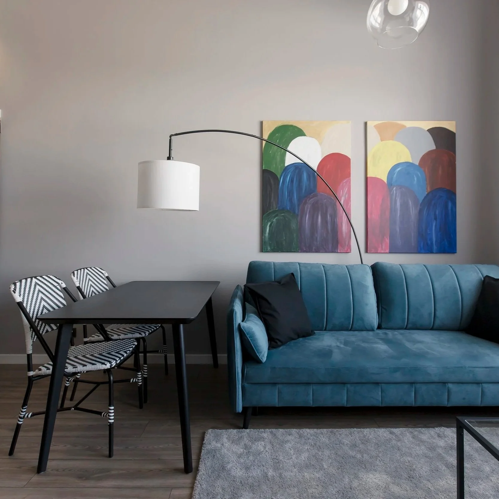 Contemporary living room with a blue velvet sofa, abstract colorful paintings on the wall, a modern arc floor lamp, a black dining table, four black and white patterned chairs, and a beige area rug on dark hardwood flooring.