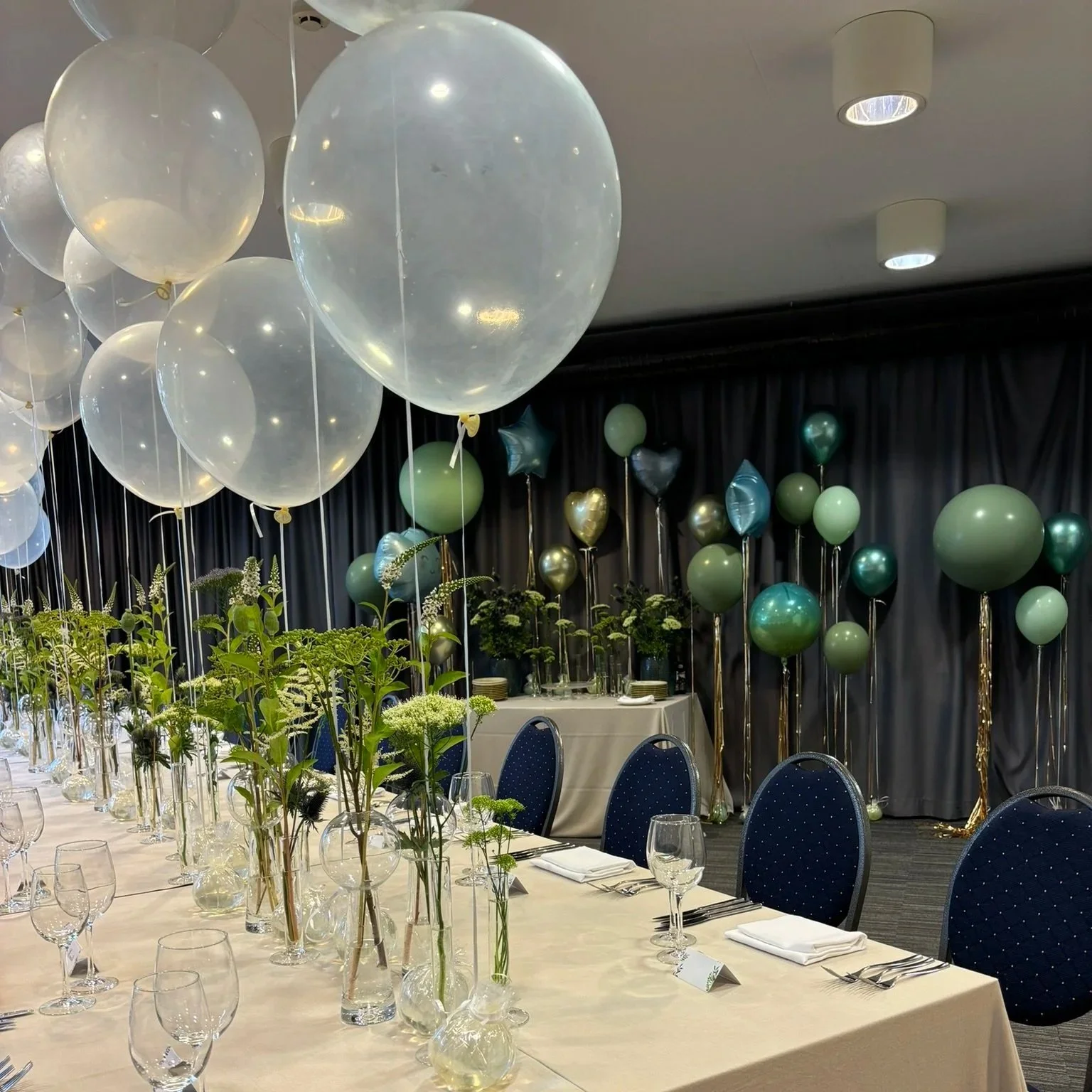 Decorated banquet hall with white tablecloths, floral centerpieces, and an array of white, green, and metallic balloons on a dark backdrop.