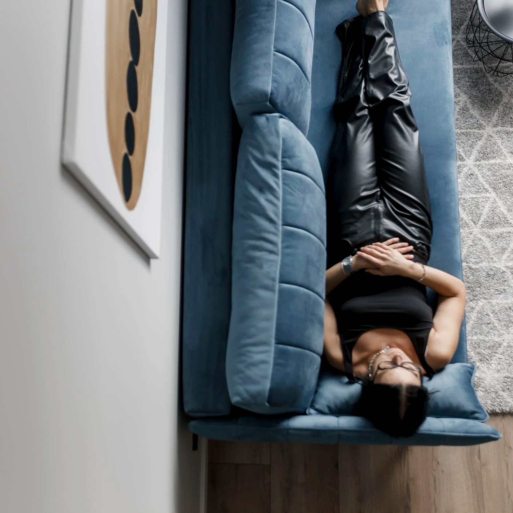 A woman lies on a blue couch with her hands resting on her forehead, wearing glasses, black clothing, and jewelry in a modern living room with gray carpet and abstract artwork on the wall.