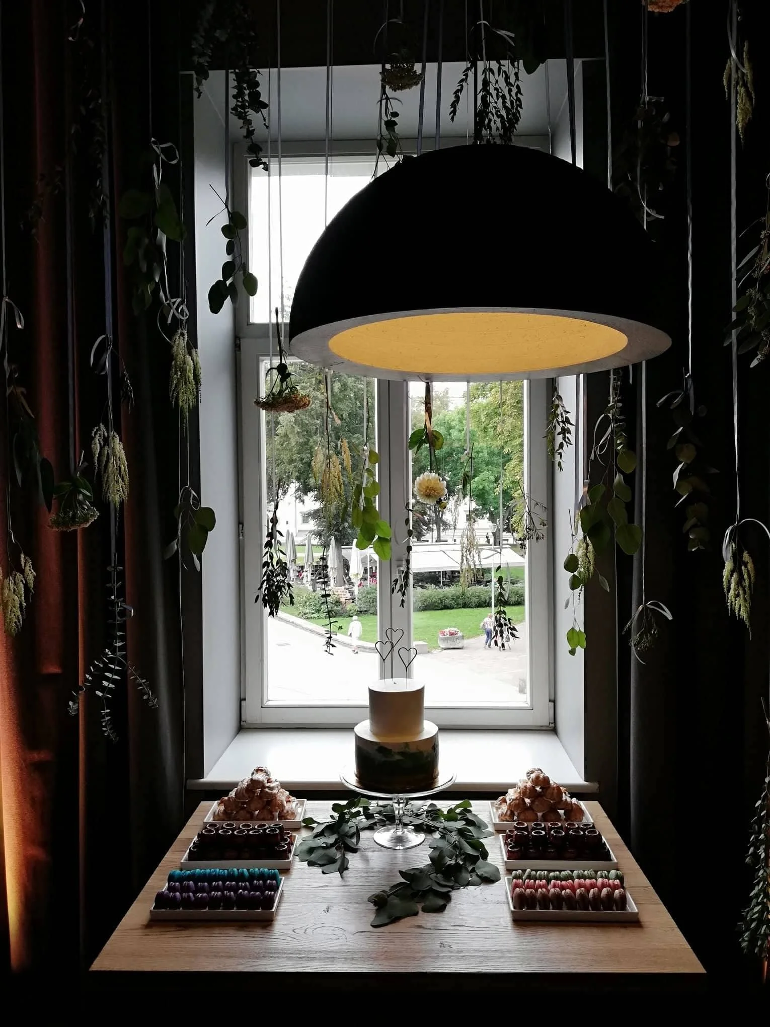 Table with macarons and chocolates, decorative greenery, potted plant, large window with view of park, hanging plants, and a large pendant light in a room overlooking a park.