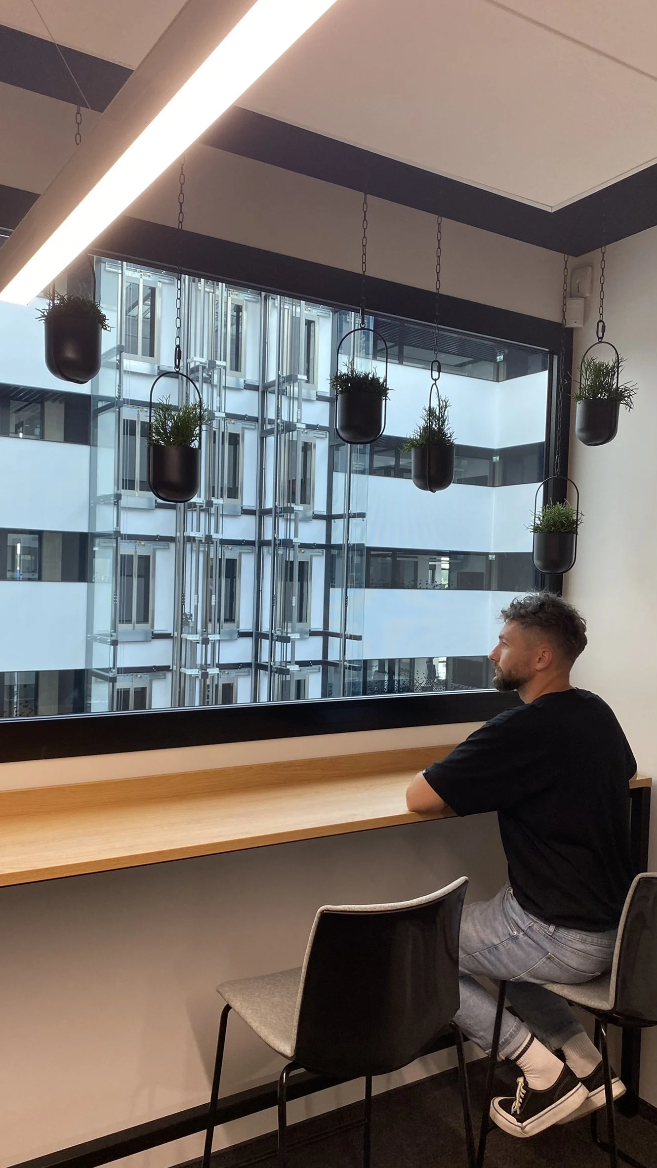 Man sitting by a wooden counter in a modern cafe or office, looking out the window with hanging planters with green plants above him, a large window revealing a multi-floor building exterior with glass and metal elements.