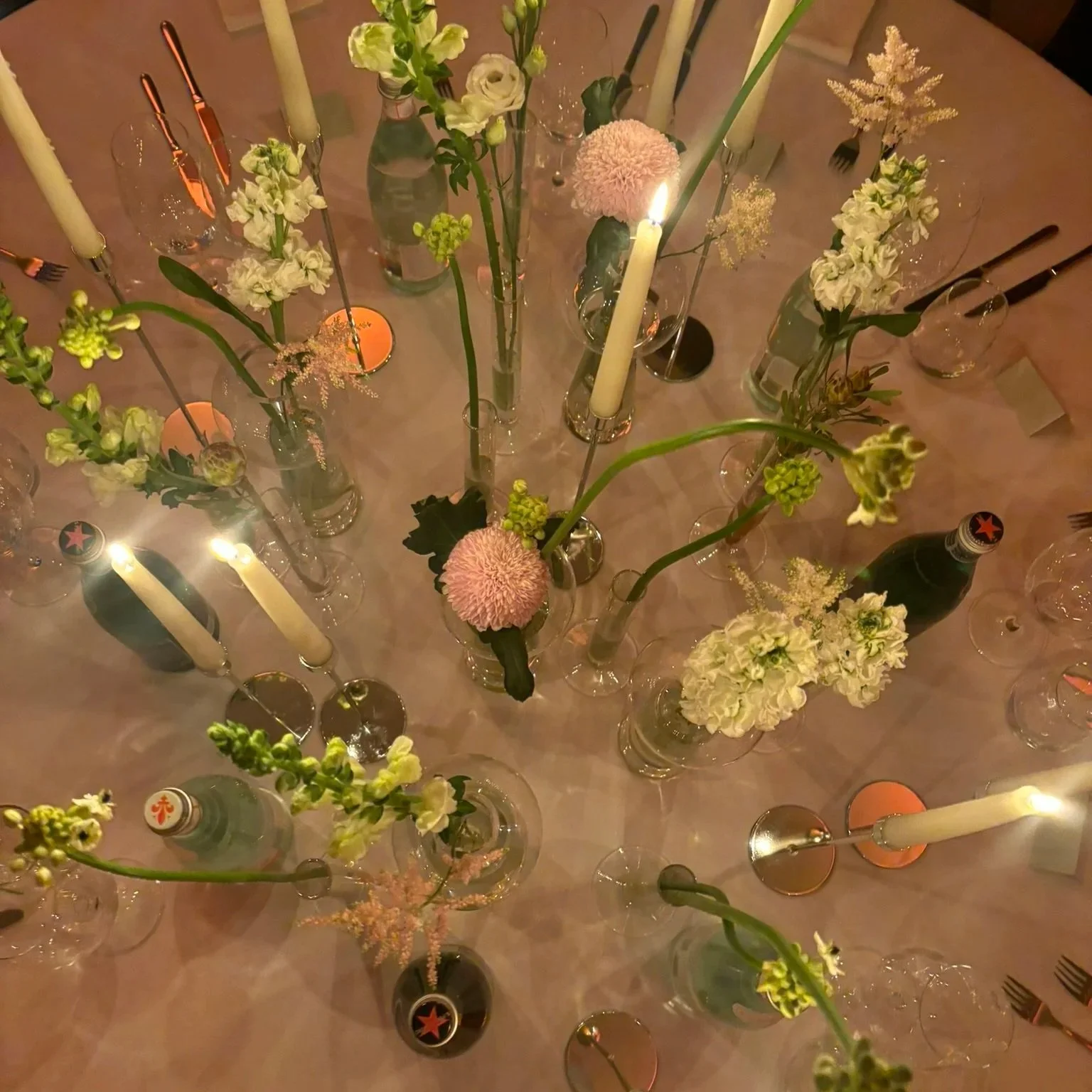 A dinner table decorated with a neutral tablecloth, featuring multiple glass vases filled with various flowers like white and pink hydrangeas, pink chrysanthemums, and other white blooms. Several lit candles in tall holders and tea lights are also on the table, along with wine glasses, some containing wine or water, and black-handled forks and knives.