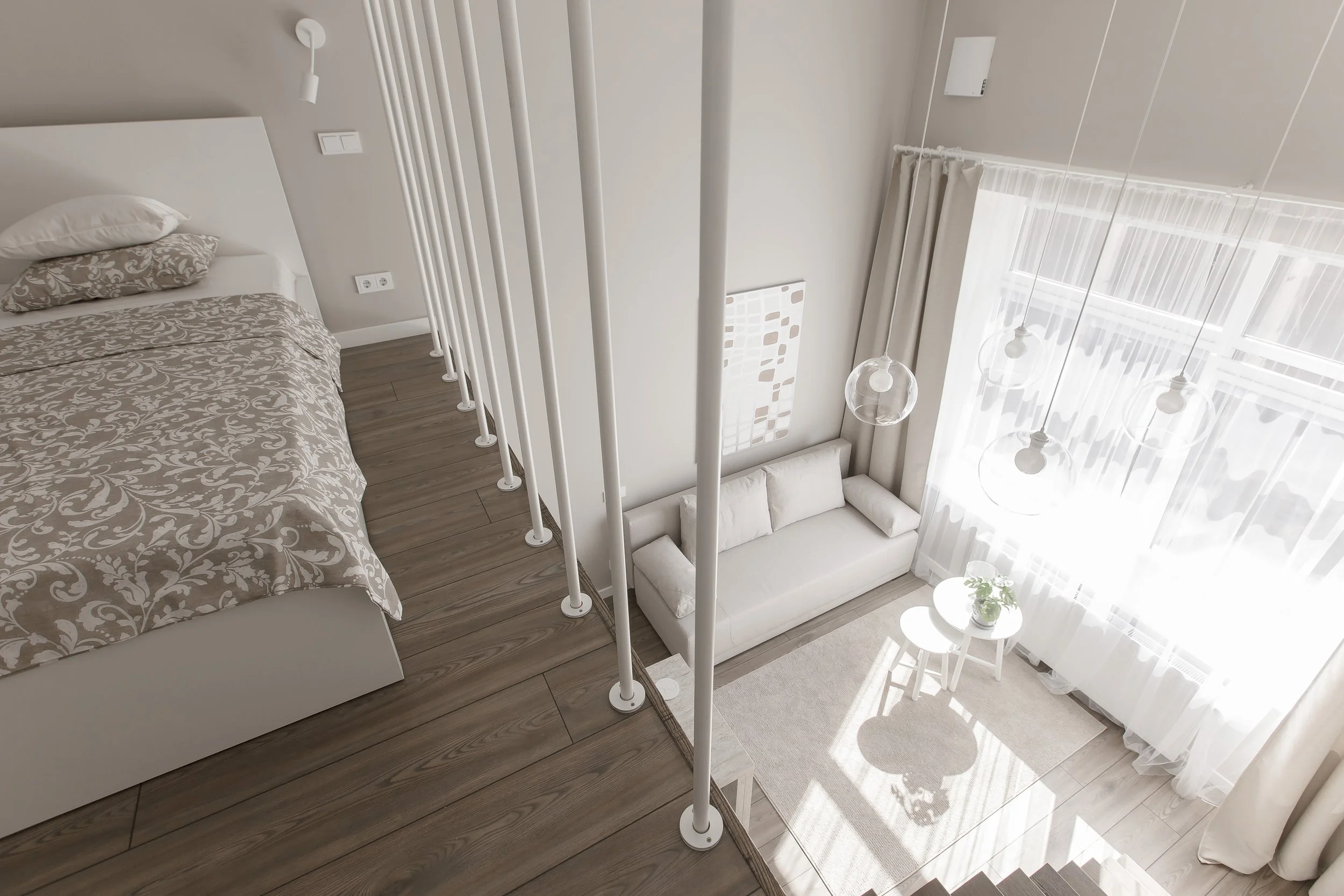 The image shows an interior view of a modern, sunlit living space with a staircase railing separating a bedroom with a bed and patterned bedding from a lower sitting area. The sitting area has a white couch, a round white side table with a plant, and large windows with sheer curtains allowing natural light to fill the room.