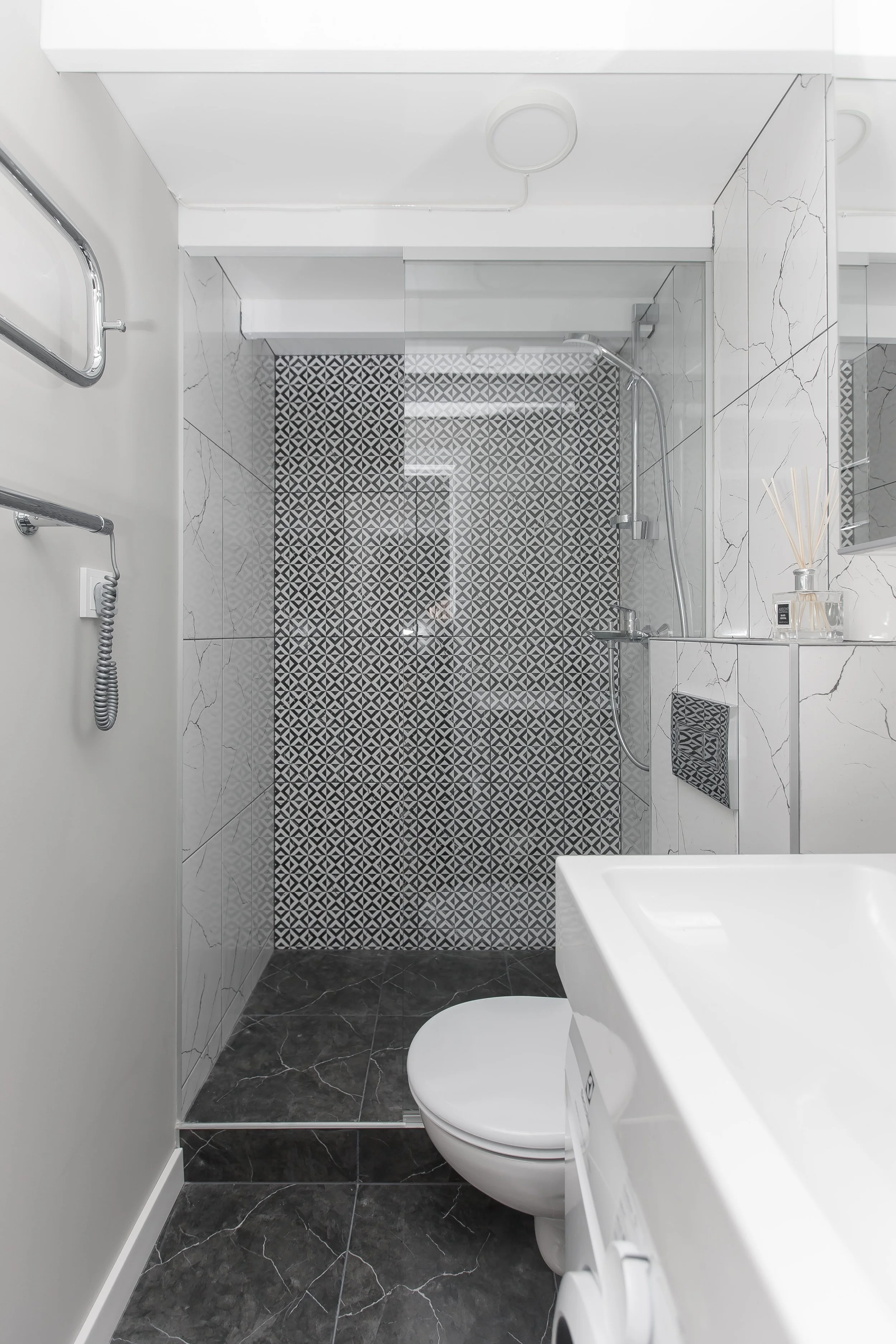 Modern bathroom with a walk-in shower, black patterned tile floor, white marble walls, a white toilet, and a white bathtub, with minimalistic decor and accessories.