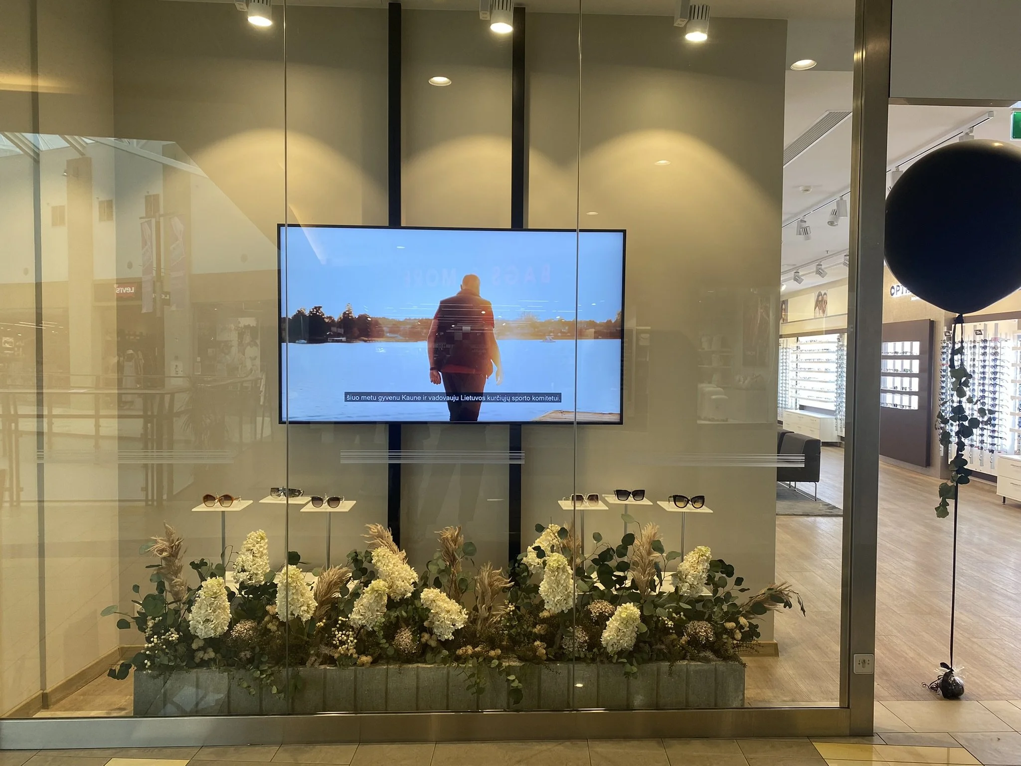 Store window display with flower arrangement at the bottom, a flat-screen TV showing a man in front of a lake, and a black balloon on the right side. The background reveals the interior of a shopping mall with lighting, signage, and shelves.
