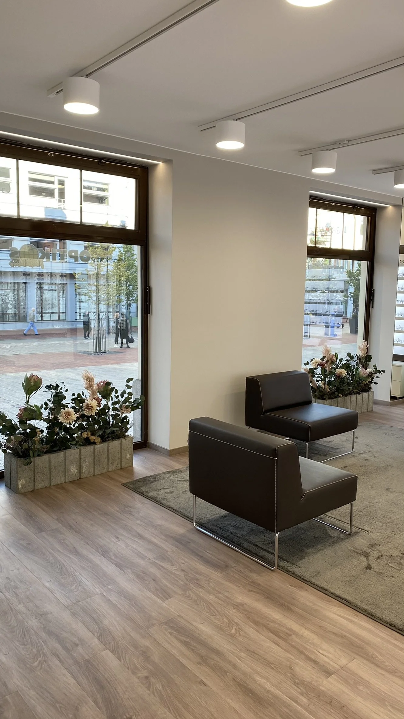 A modern indoor waiting area with two black leather armchairs, a beige area rug, and two flower planters with pink and white flowers near large windows. Outside, a city street with pedestrians and buildings is visible.