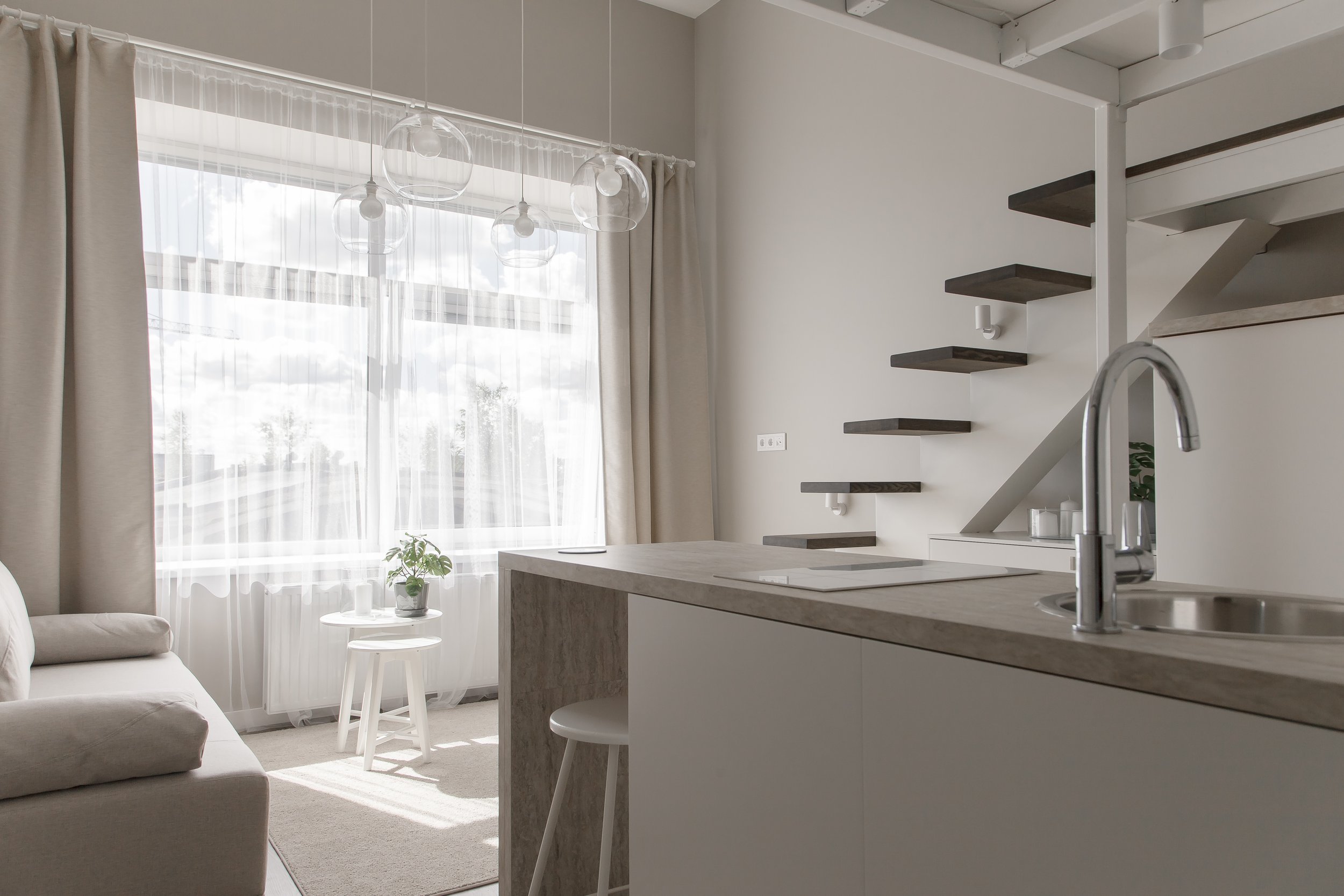 Bright living space with large window, beige curtains, white sheer drapes, a beige sofa, small white side table with a potted plant, hanging glass pendant lights, open kitchen with a light stone countertop, staircase with floating dark wood steps, and a white wall with electrical outlets.