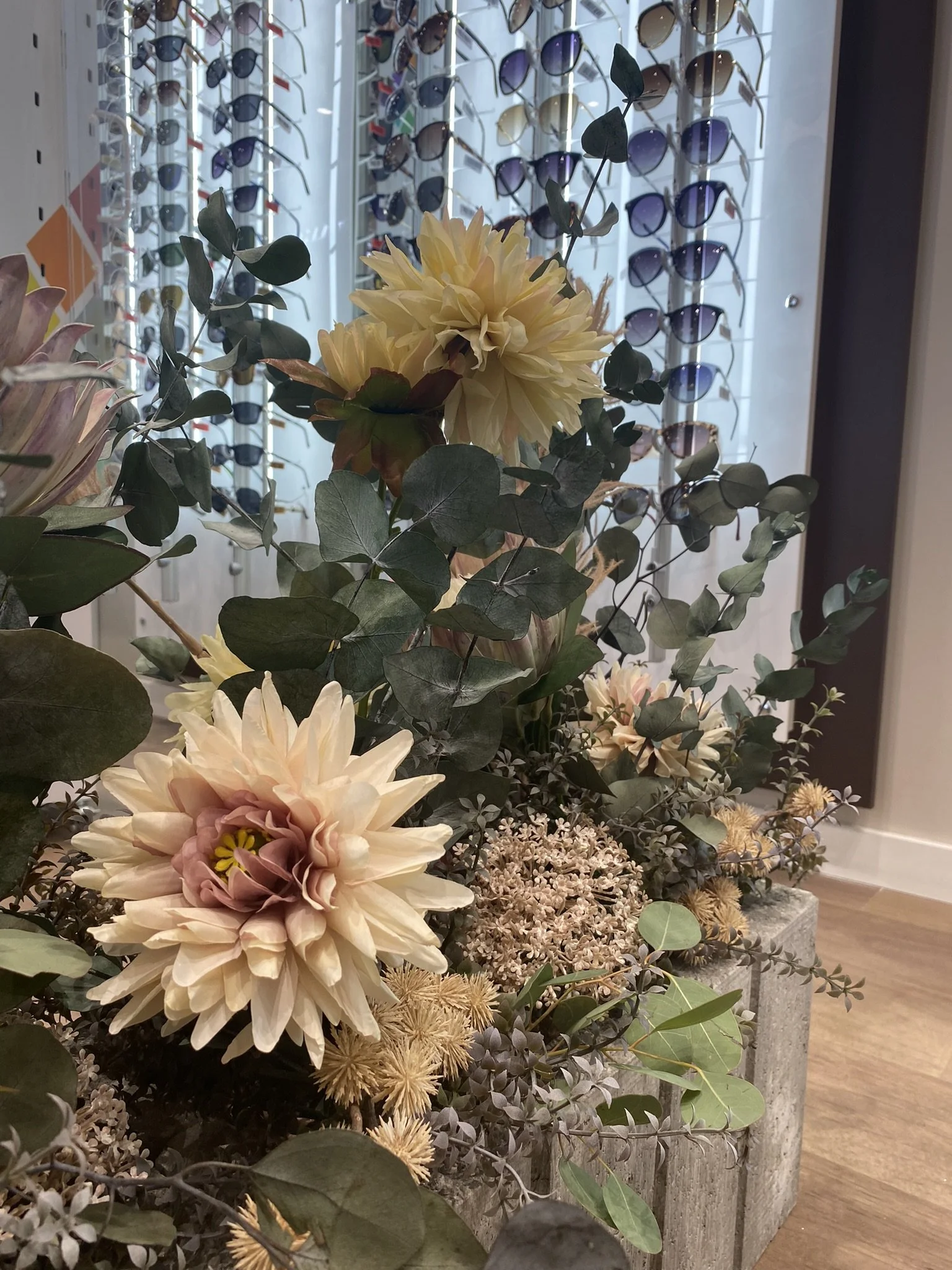 A display of various artificial flowers and greenery arranged in a decorative container near sunglasses displayed on a wall in the background.