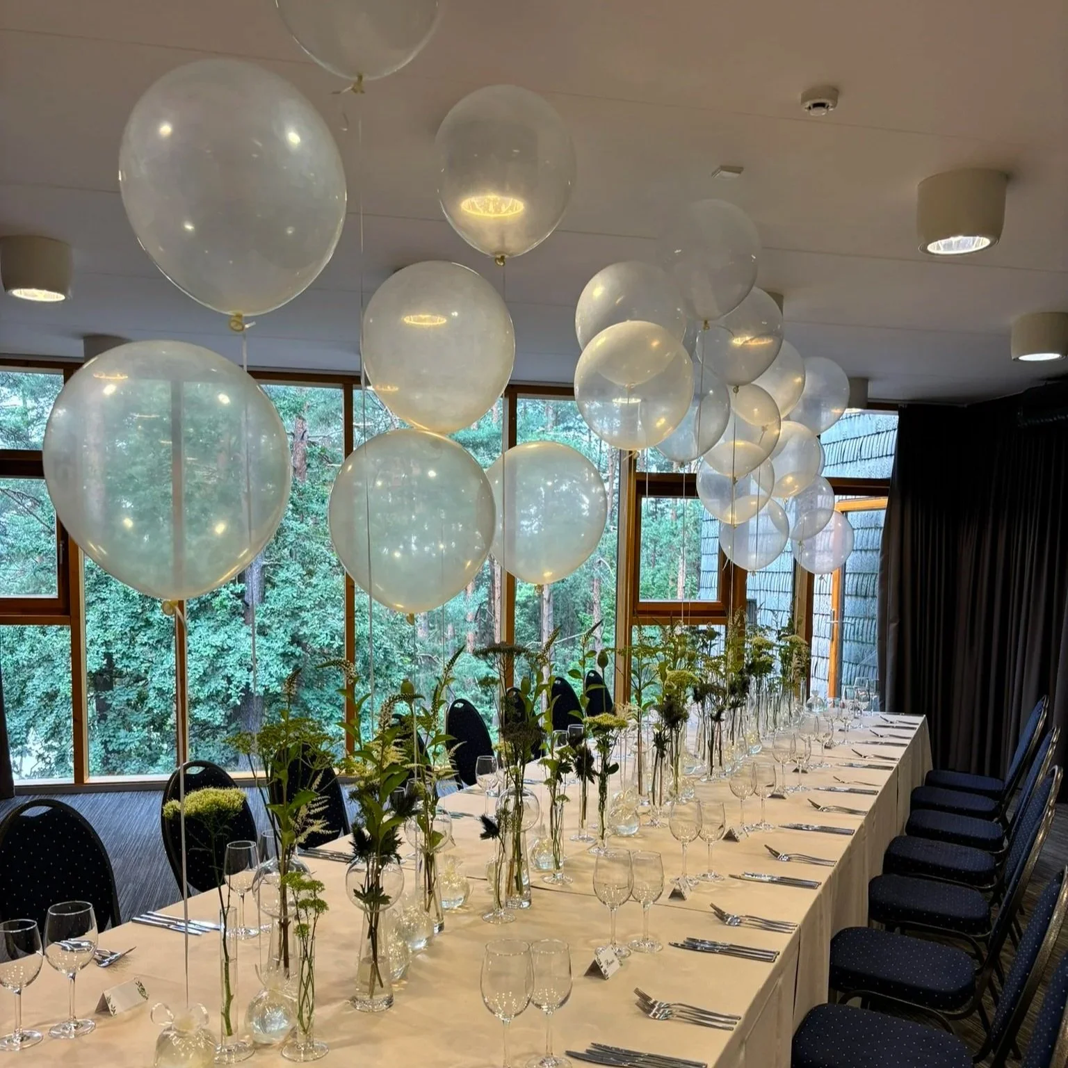 Long dining table set for a celebration, decorated with tall vases of flowers and surrounded by black chairs, with white balloons hanging from the ceiling and large windows with views of trees.