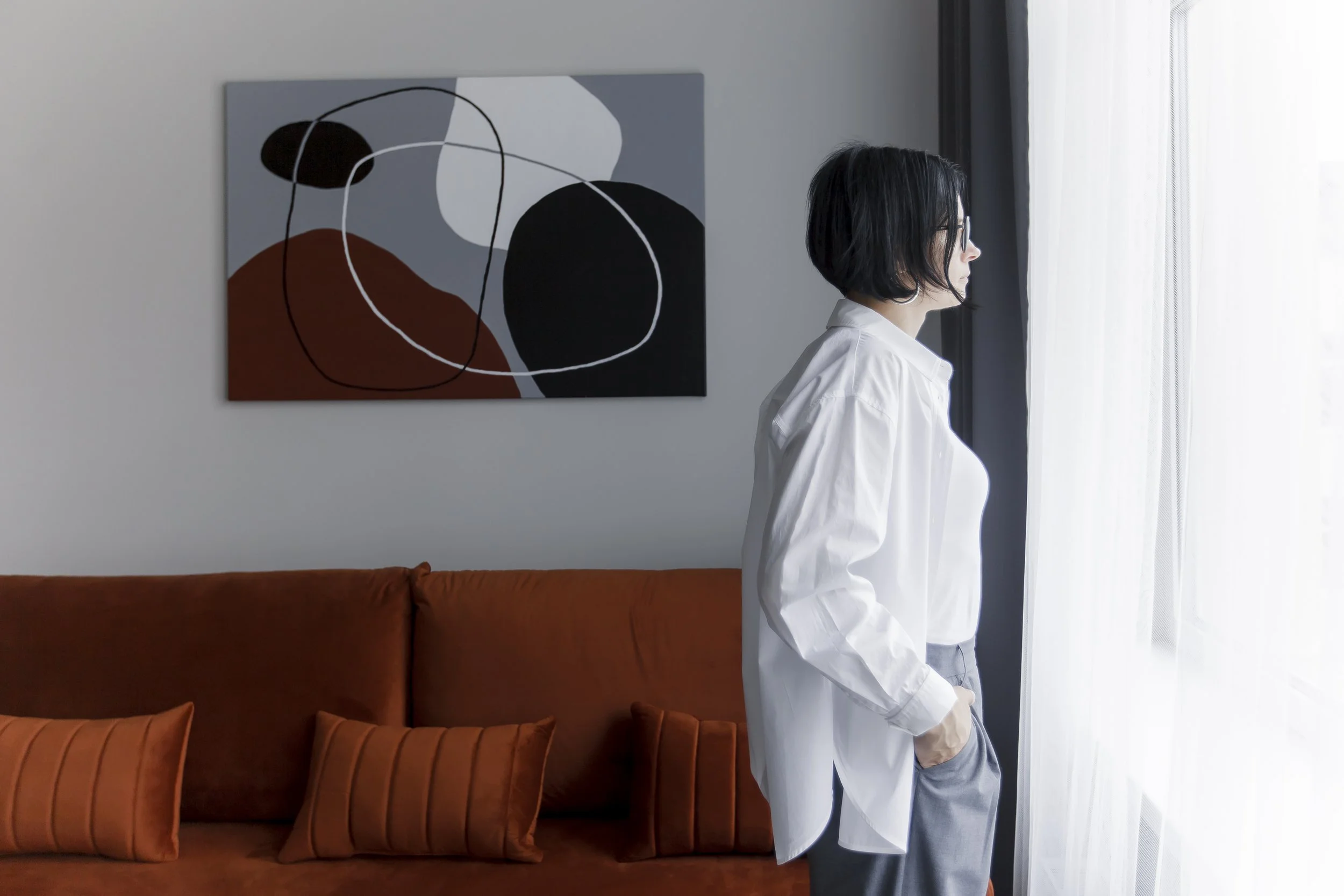 A woman with short black hair, wearing glasses and a white shirt, standing sideways by a window with sheer curtains, in a living room with a brown sofa and an abstract painting on the wall.