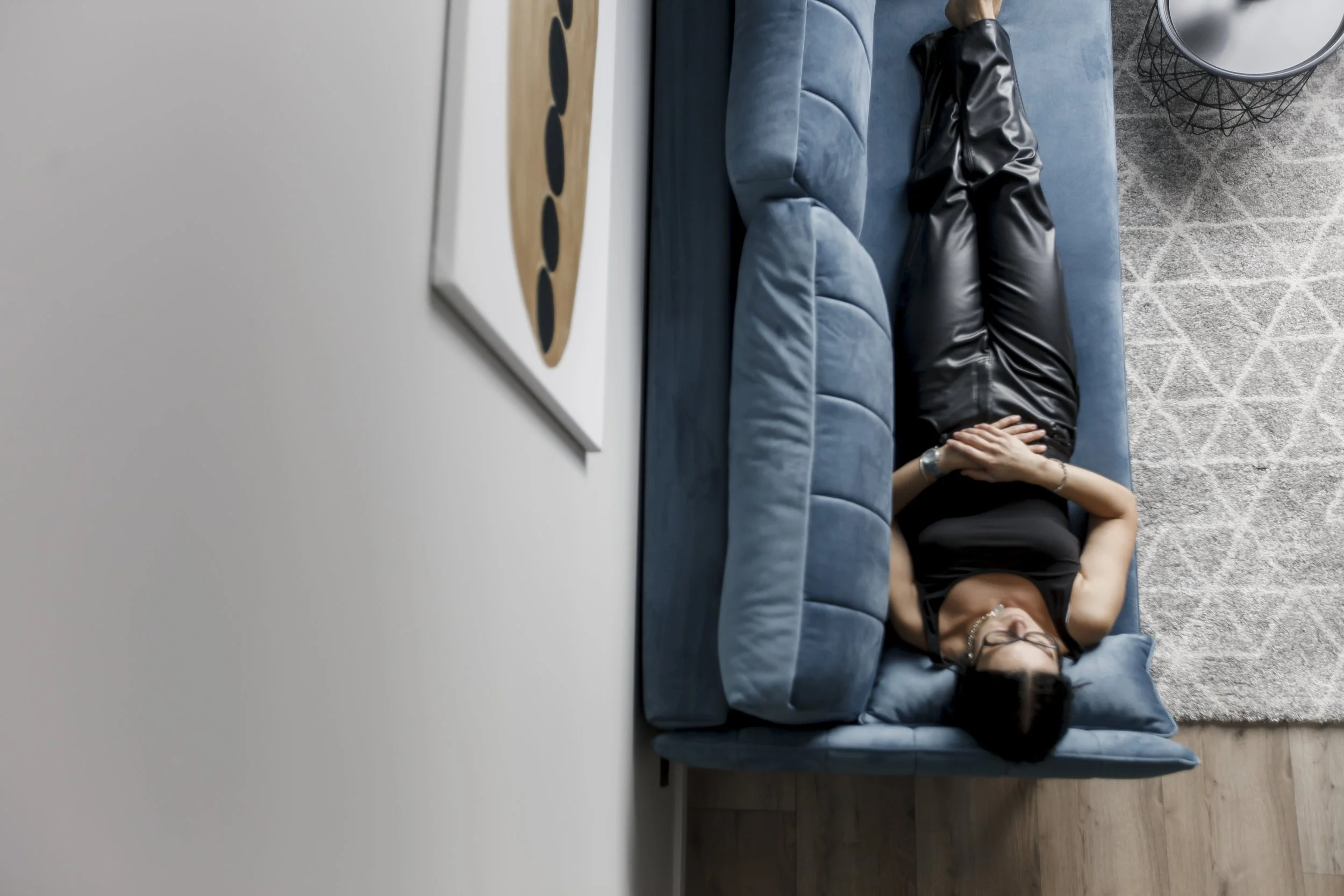 A woman is lying on a blue velvet couch in a modern living room, with her hands holding her face, while wearing a black shirt and black leather pants. The room has a gray patterned rug, a round side table with a wireframe base, and a framed, abstract