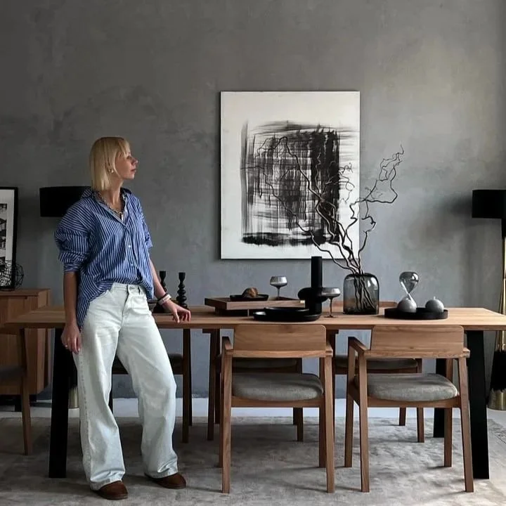 A woman with blonde hair wearing a blue and white striped shirt and white pants, standing next to a wooden dining table in a modern, minimalistic room with gray walls, black and white artwork, and decorative vases on the table.