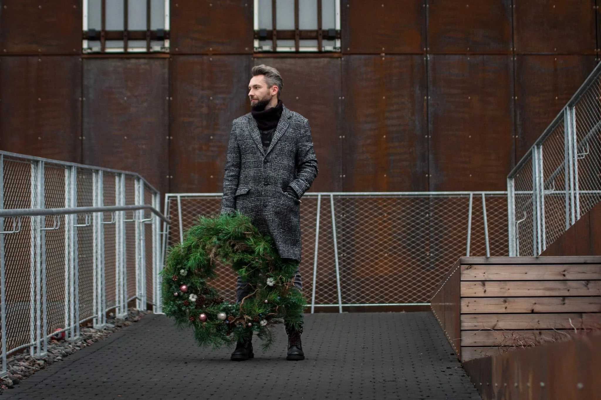 A man with a beard and styled hair walking outside on a paved pathway, holding a Christmas tree decorated with ornaments. He is wearing a checkered coat and a black scarf, with a modern building with large wooden panels in the background.
