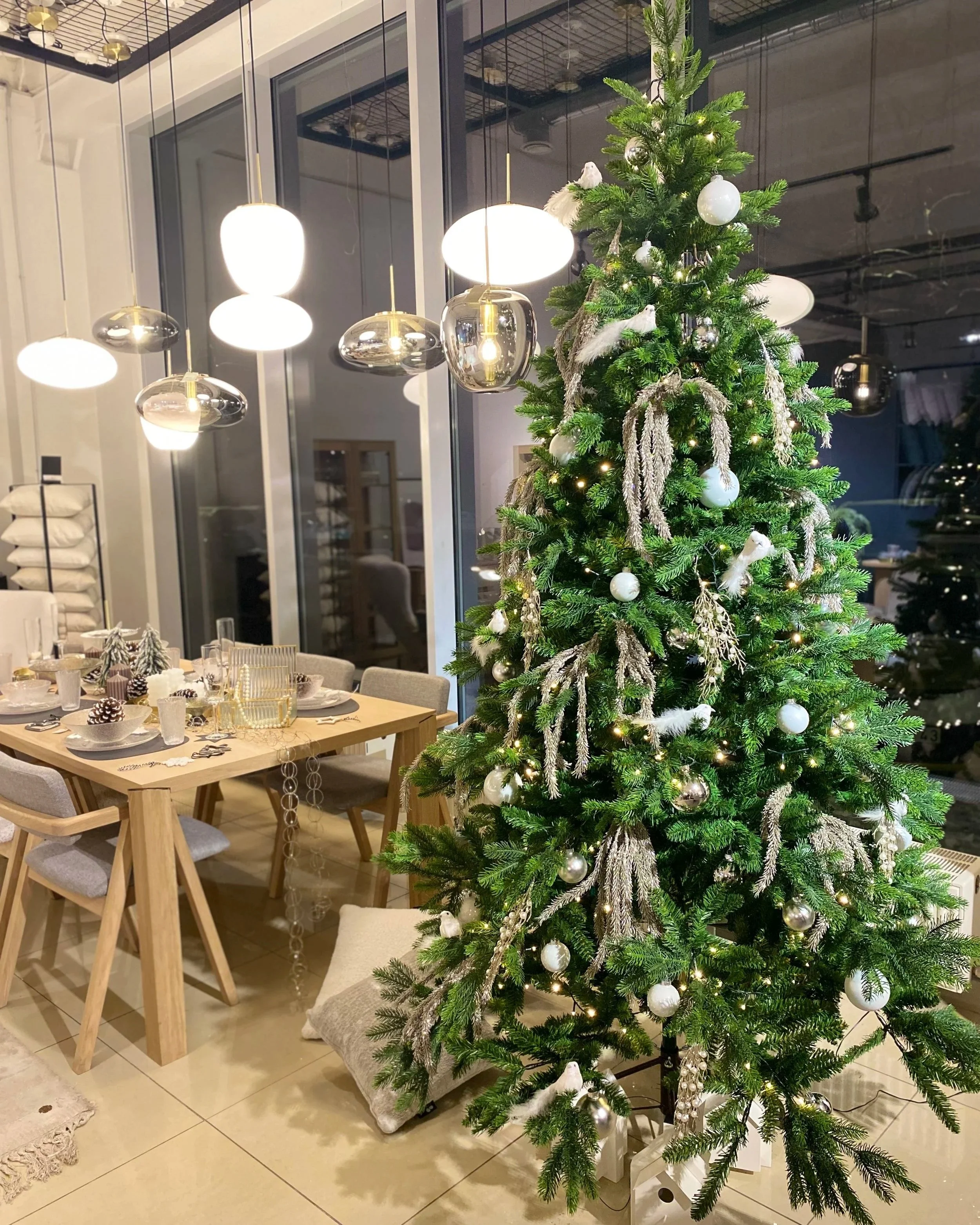A decorated Christmas tree indoors beside a dining table with holiday decorations, including pinecones, candles, and ornaments, under modern hanging lights.