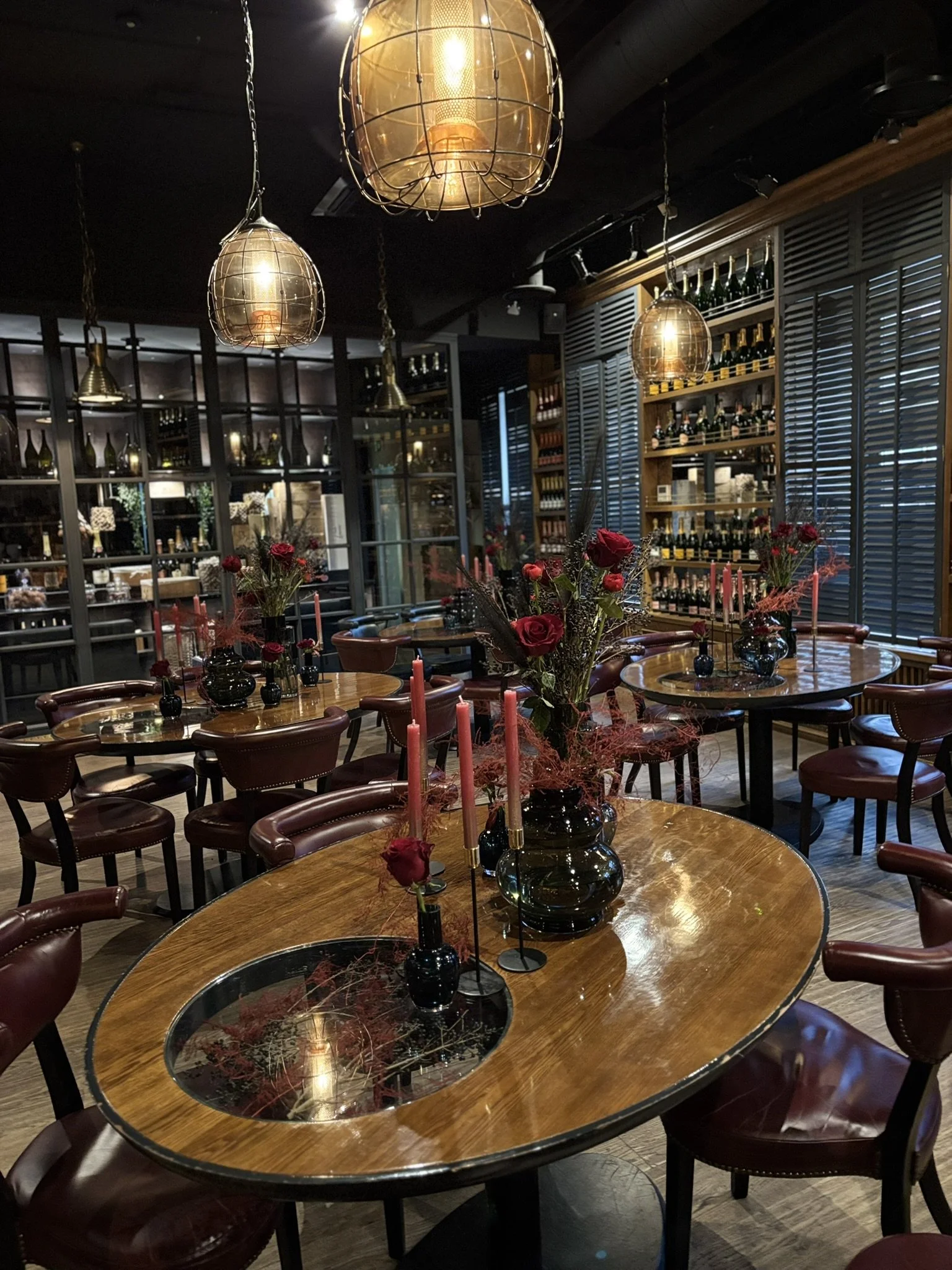 Interior of a restaurant with wooden tables, burgundy chairs, floral centerpieces, and warm lighting from pendant lamps and candle holders.
