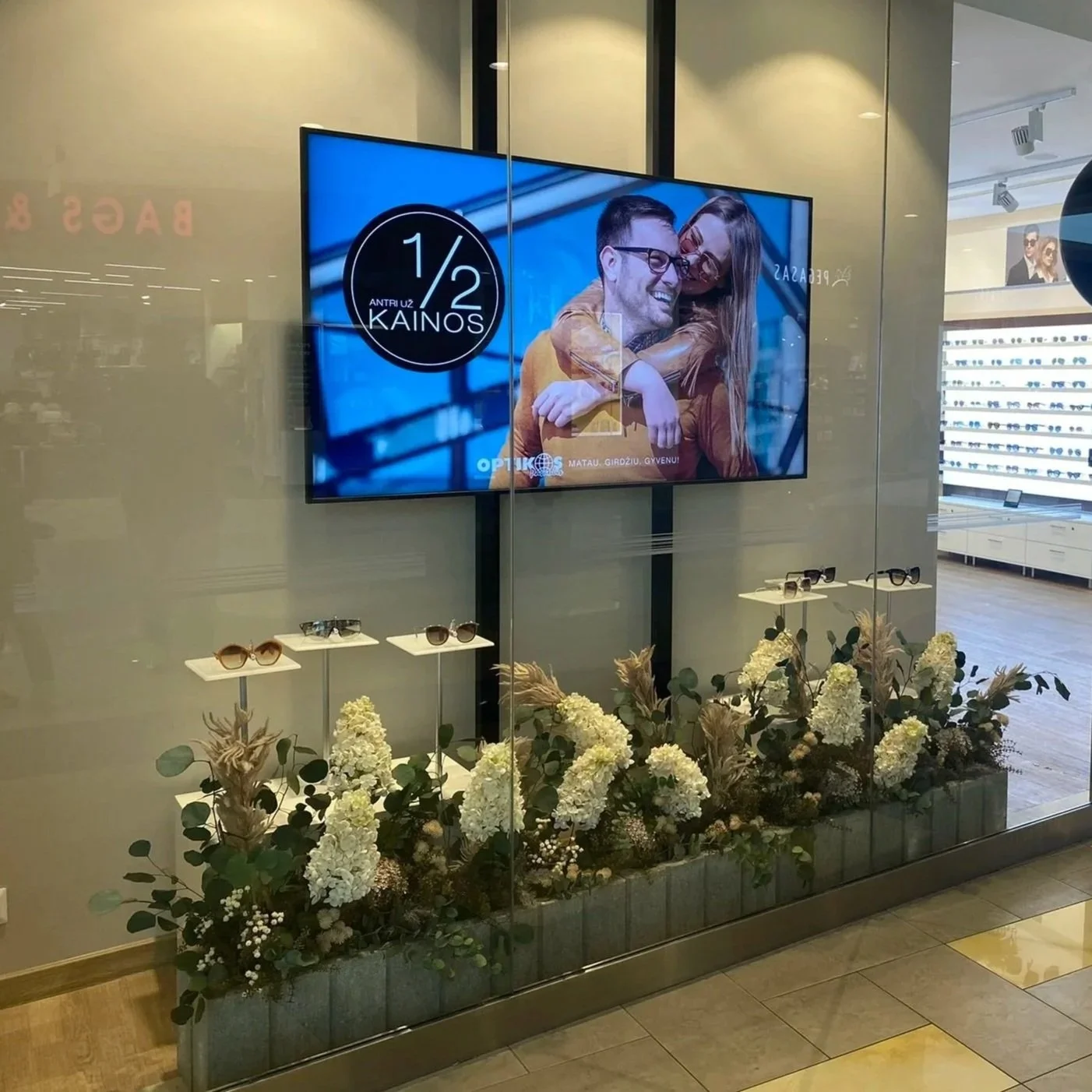 A store window display with a large electronic advertising screen promoting sunglasses with a 50% discount. The screen shows a couple smiling, embracing, and wearing sunglasses. Below the screen, there are shelves with several pairs of sunglasses, and a flower arrangement with white and beige flowers in front of the display.