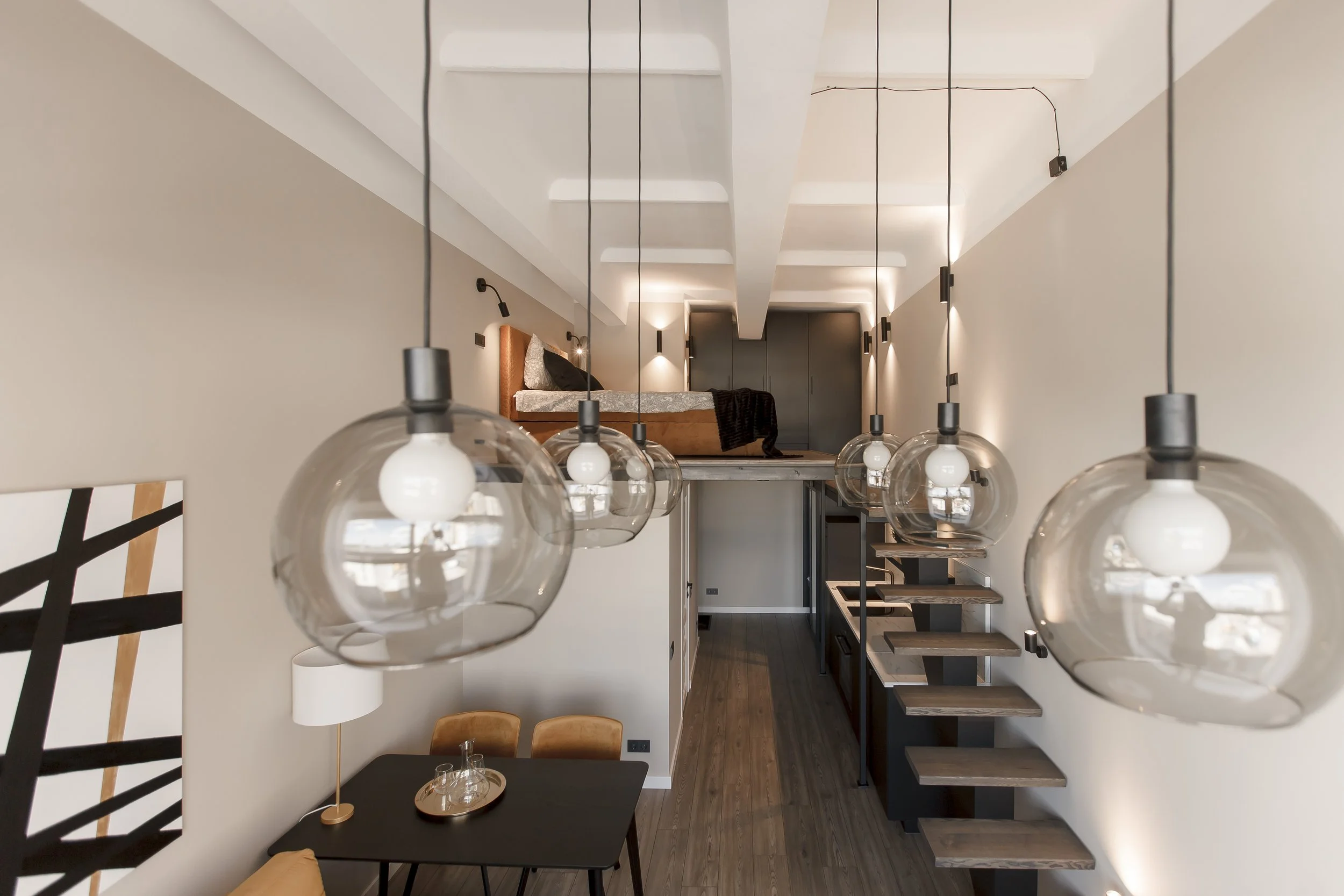 Modern dining area with a black table, wooden chairs, a floor lamp with a white shade, hanging globe pendant lights, stairs leading to a loft bedroom with a bed and dark closet, and abstract artwork on the wall.