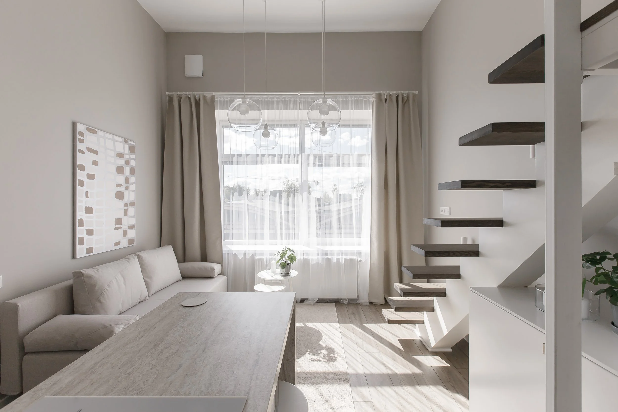 A modern living room with a white sofa, a light gray table, and floating dark wooden shelves on the wall. Large window with white curtains allows natural light to fill the space. Overhead, there are three hanging glass globe pendant lights.