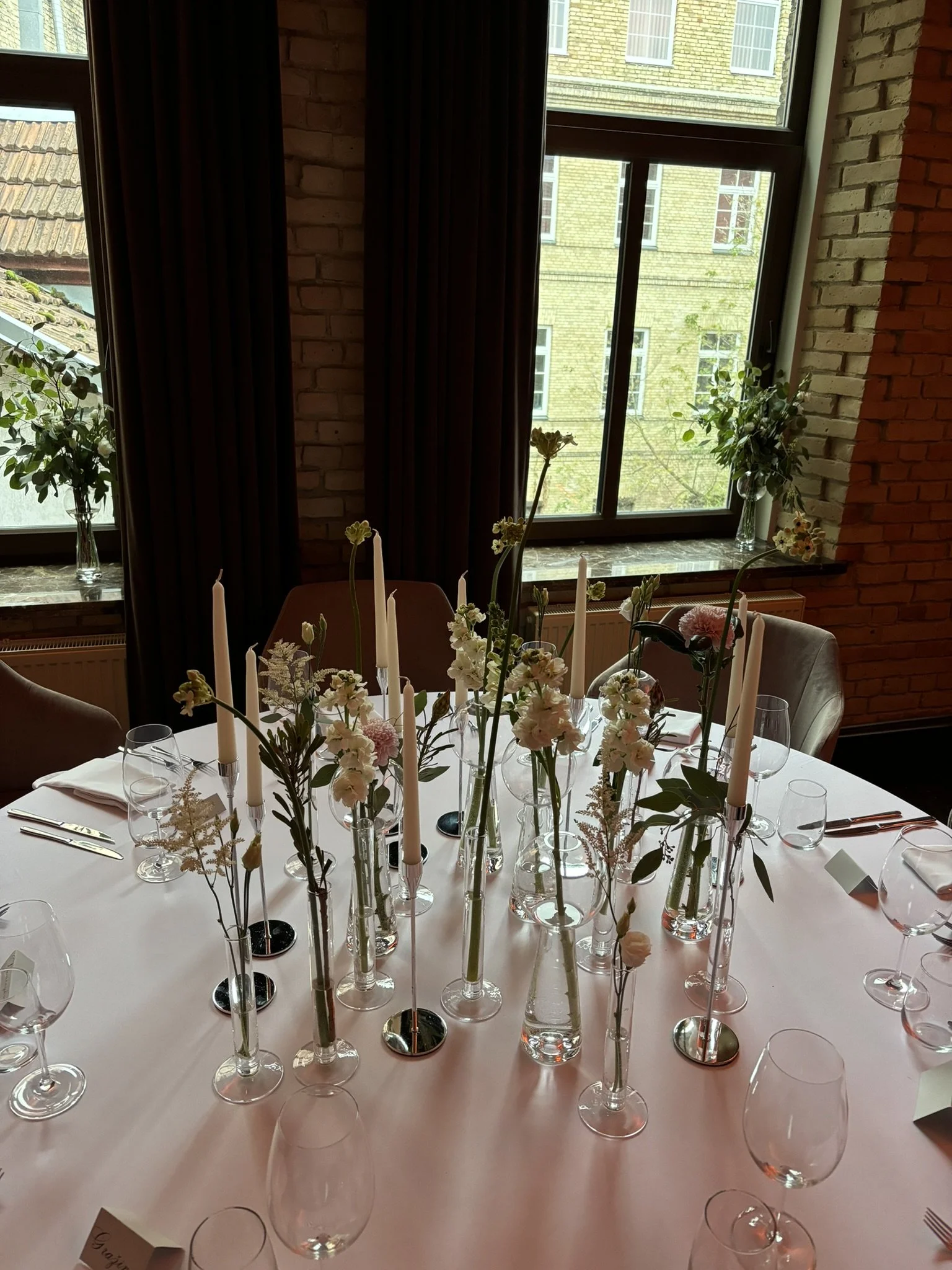 A table decorated with tall, slender vases holding white and pink flowers and long white candles, set for a formal event in a room with large windows, brick walls, and dark curtains.