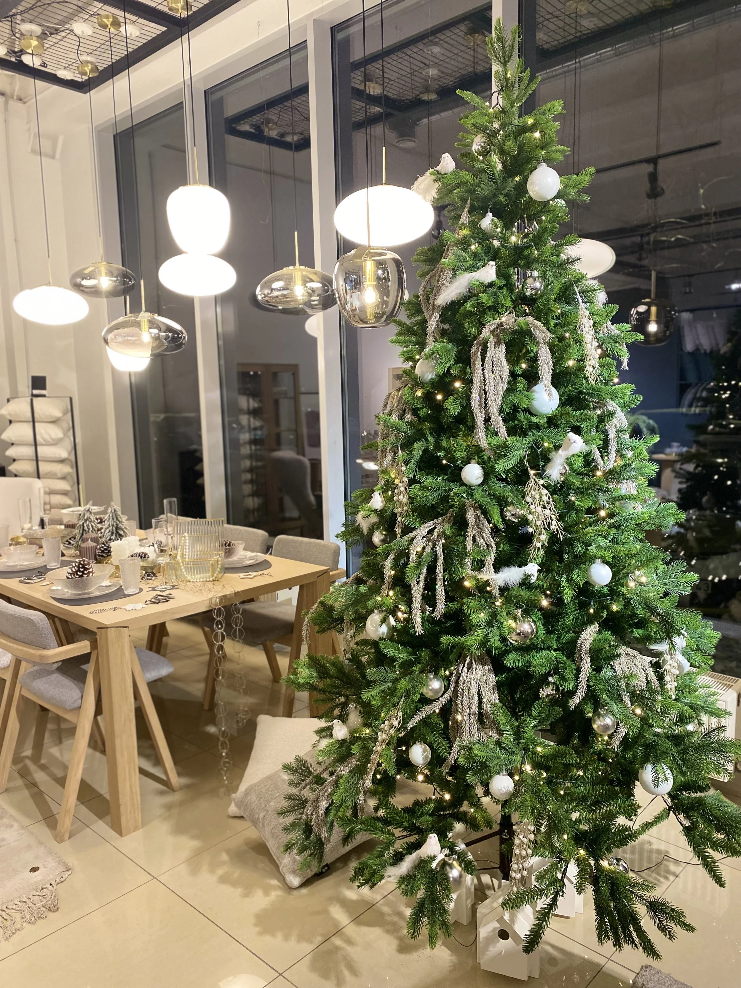A decorated Christmas tree with white and silver ornaments and lights in a modern living space near a dining table and large windows.