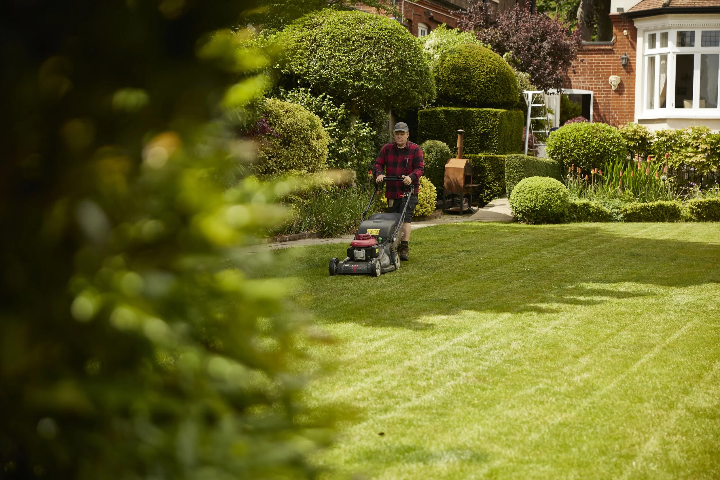 Man in red plaid shirt and cap mowing a lush green lawn in front of a red brick home with well-kept bushes and flowers.