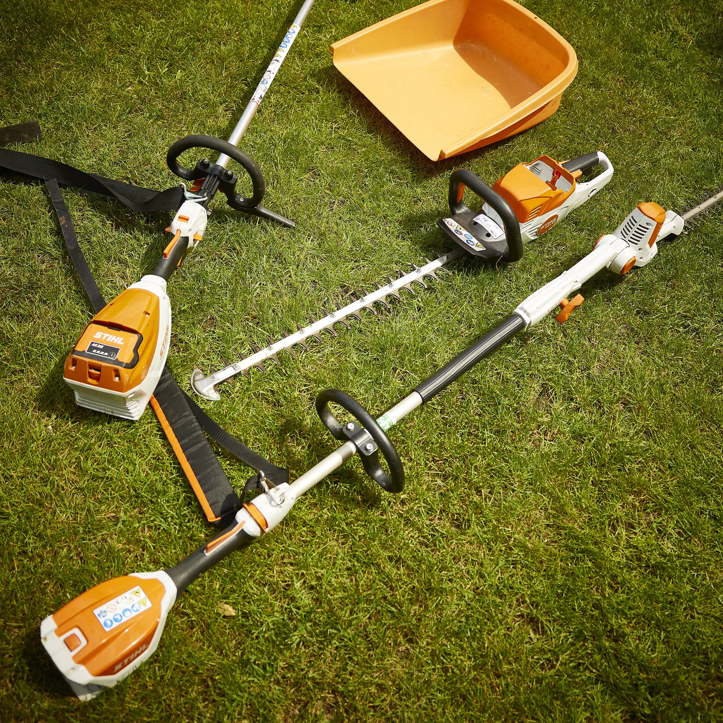 Lawn care equipment including a leaf blower, a small rake, and an orange wheelbarrow on grass.
