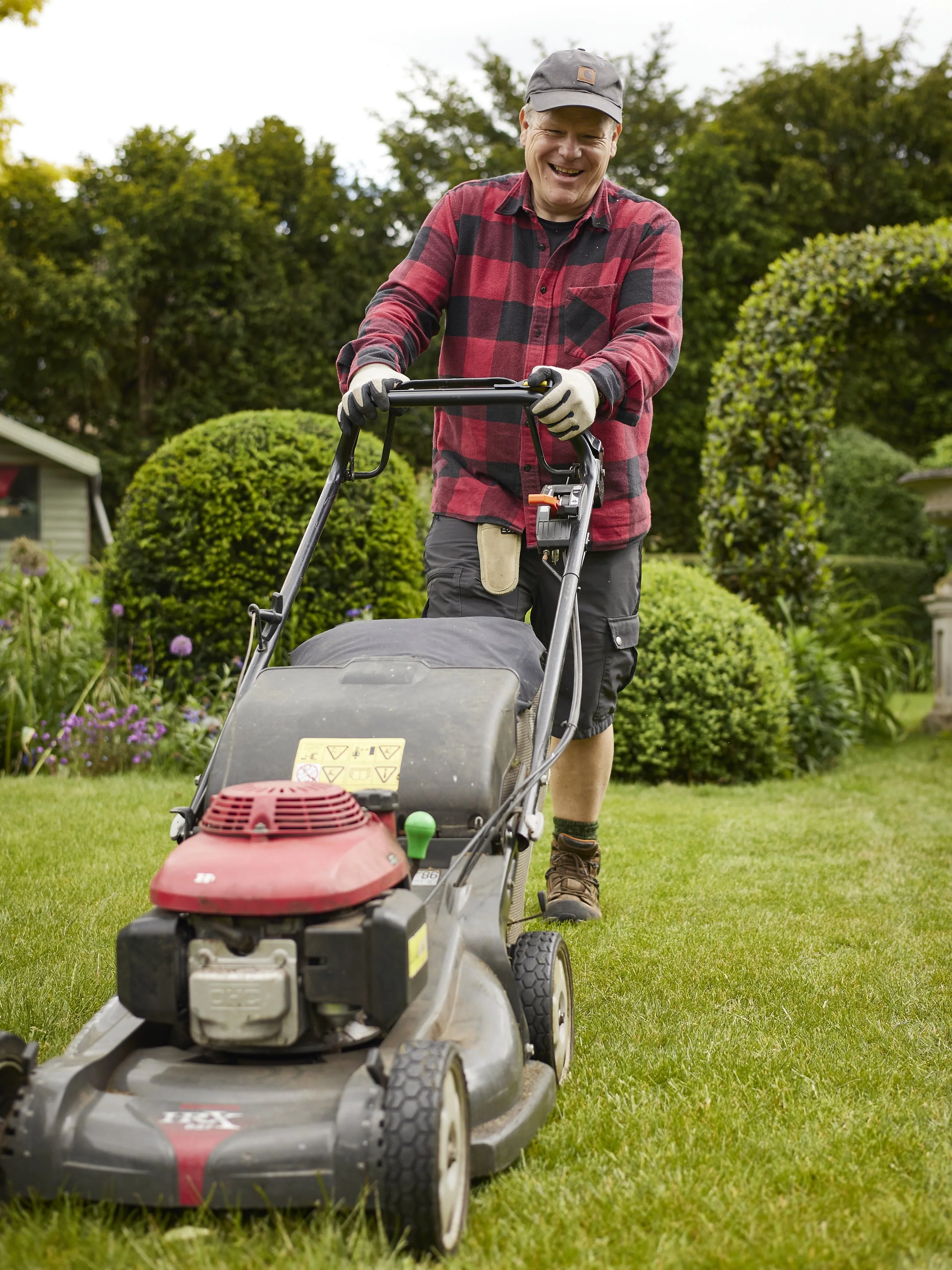 A man wearing a gray cap, red and black checked shirt, and gloves is mowing the grass with a gasoline-powered lawn mower in a lush garden with green trees and bushes.