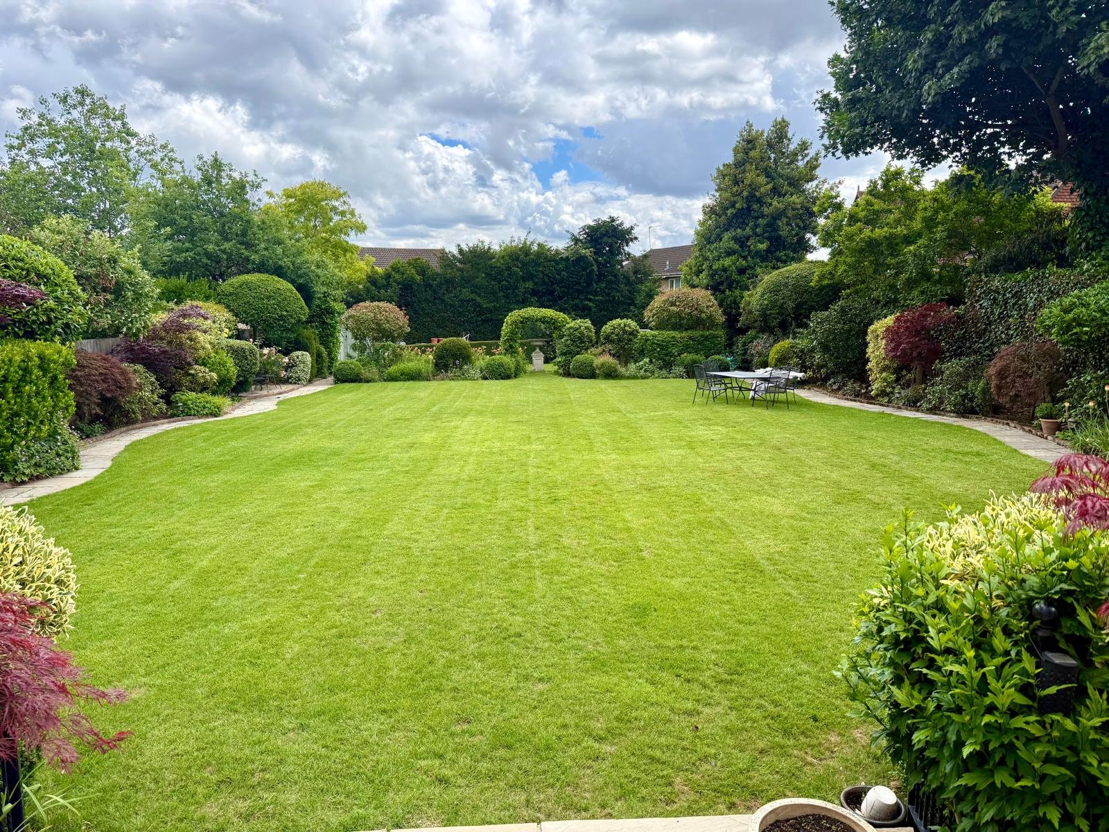 A lush green backyard with well-maintained grass, surrounded by colorful bushes and trees, with a stone pathway on either side and outdoor furniture on the right.