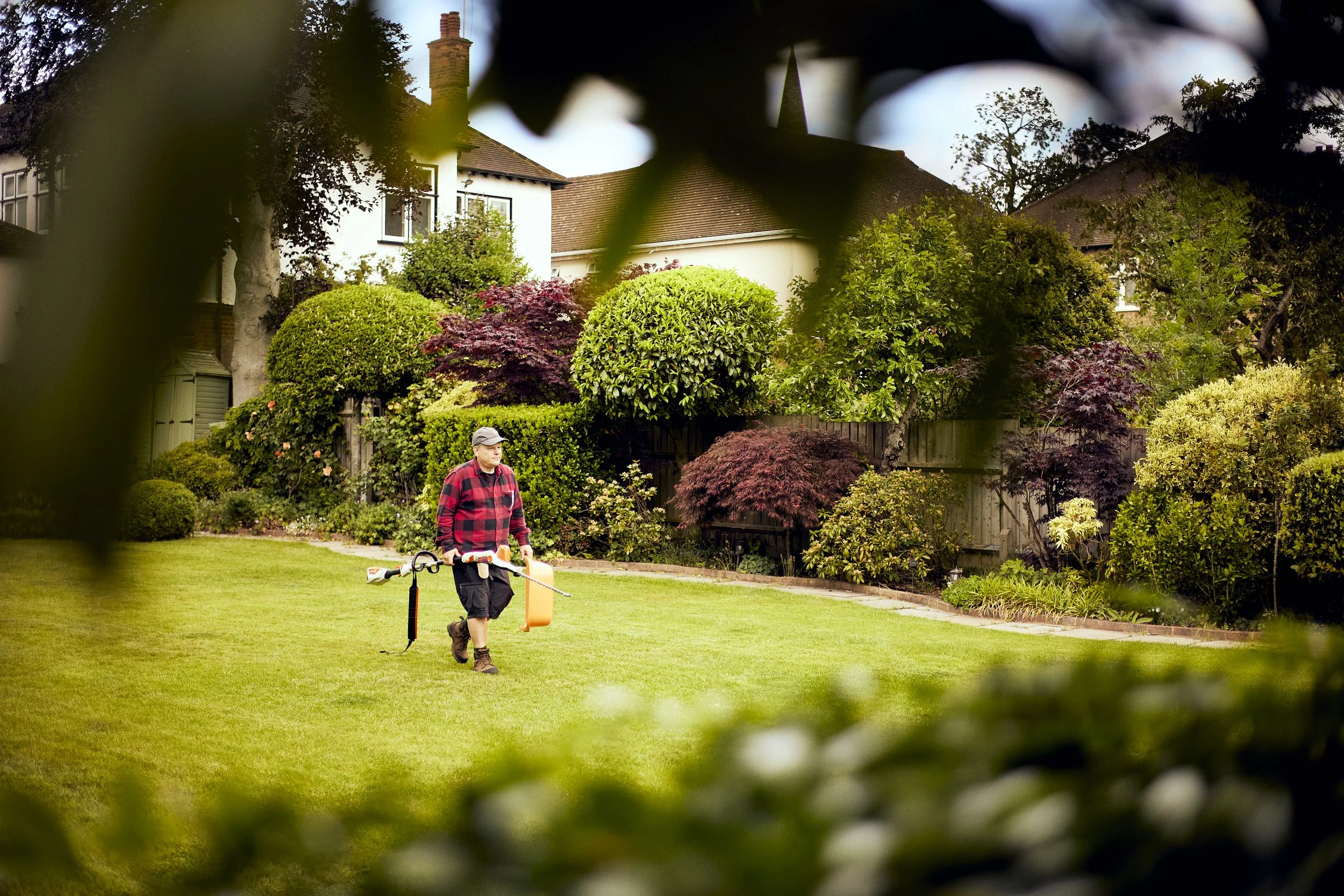 A man wearing a plaid shirt, cap, and shorts walks on a grass lawn while holding a spray device. He is surrounded by lush, well-maintained garden bushes and trees, with houses and a clear sky in the background.