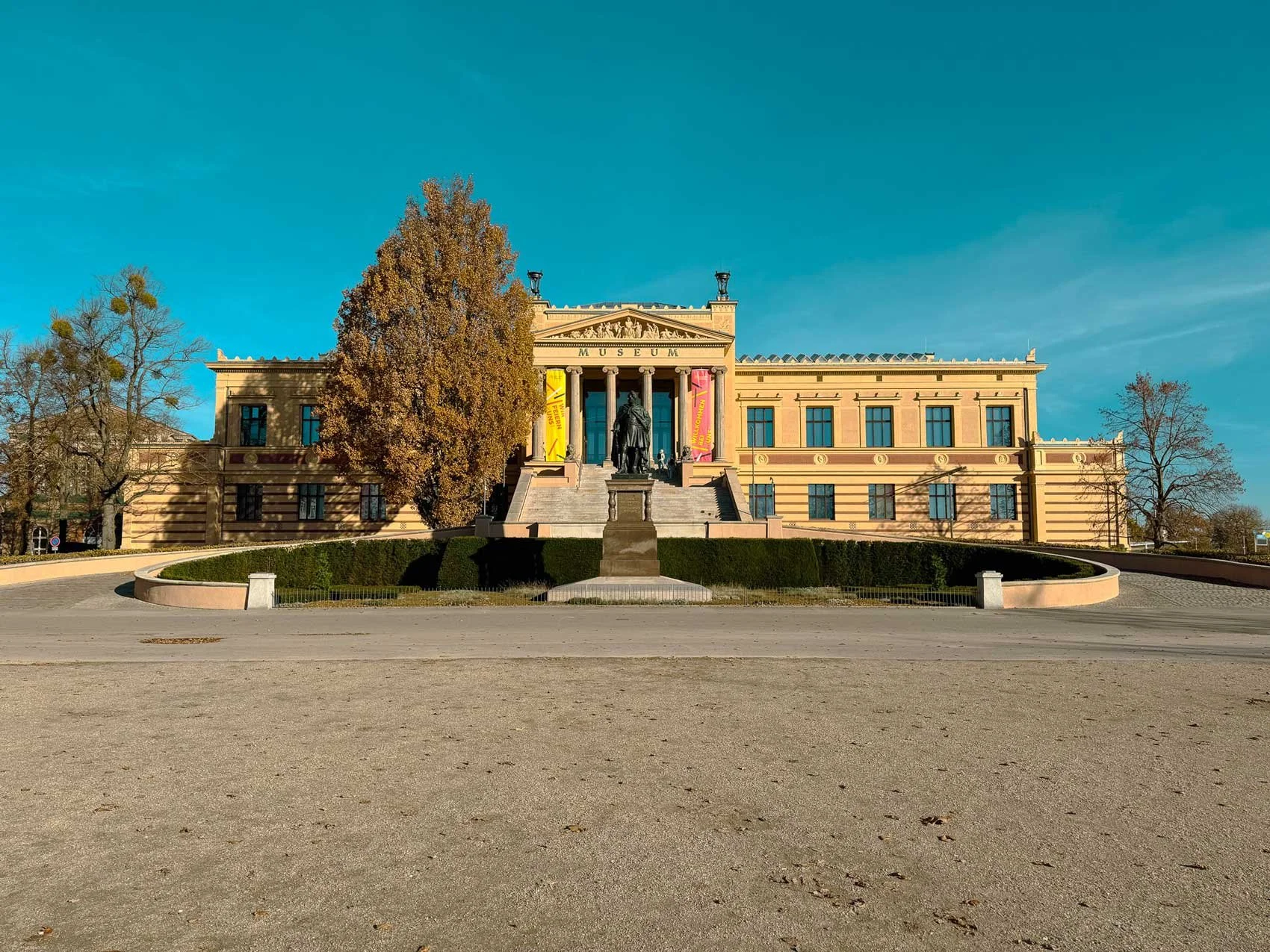 Ein klassisches, gelb gestrichenes Museum mit Säulen vor einem blauen Himmel, umgeben von Bäumen und einem gepflegten Garten mit Kalksteinmauer